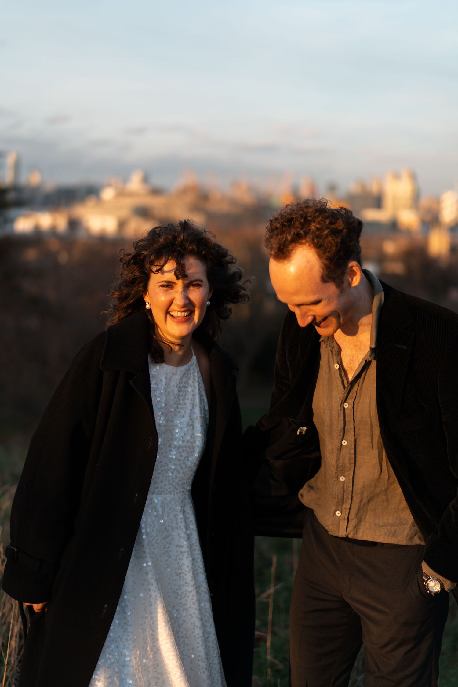 A joyful couple laughs together on a hill during golden hour with a cityscape behind them.
