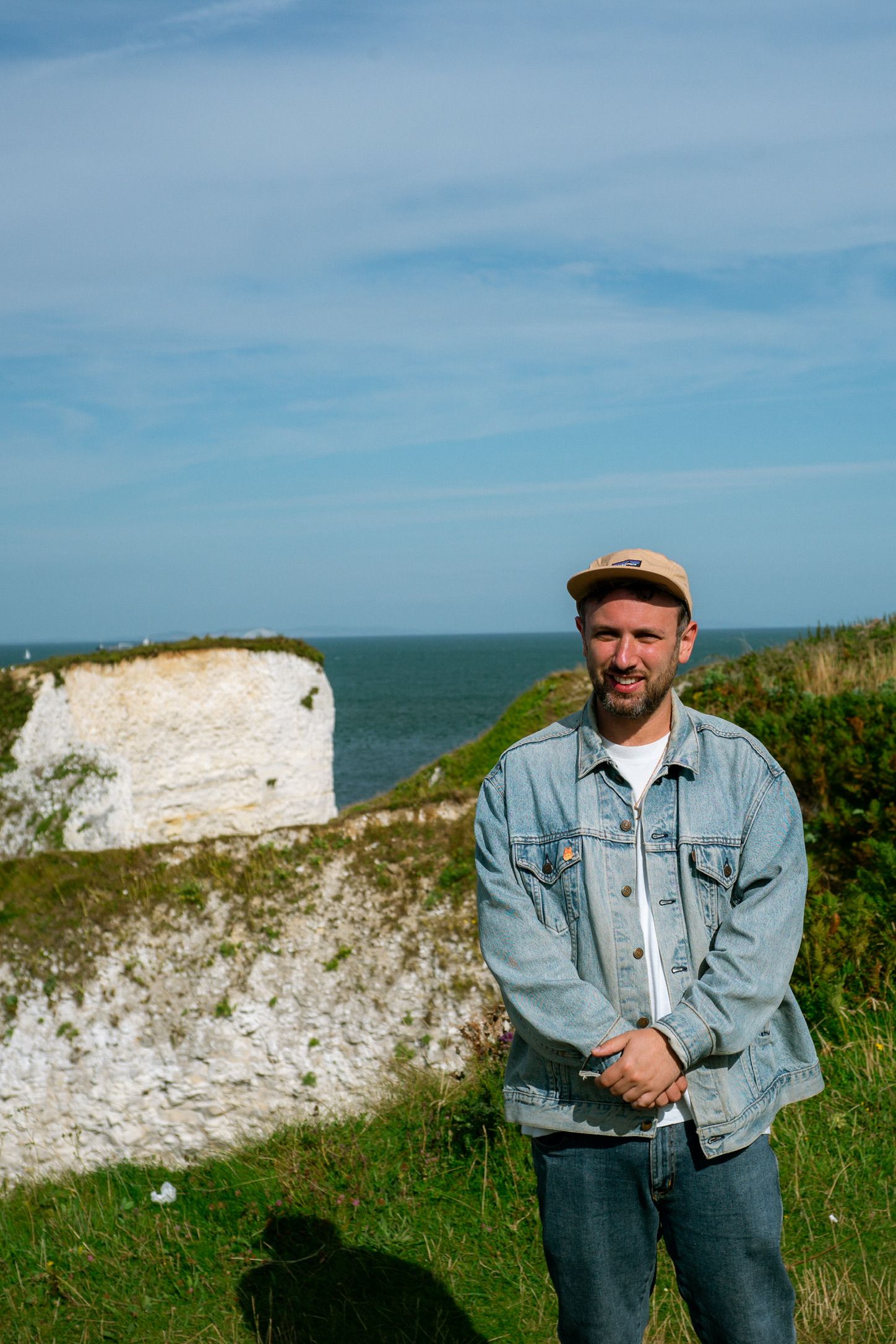 Man in denim jacket and cap stands on green cliffs above white chalk sea cliffs under a blue sky.