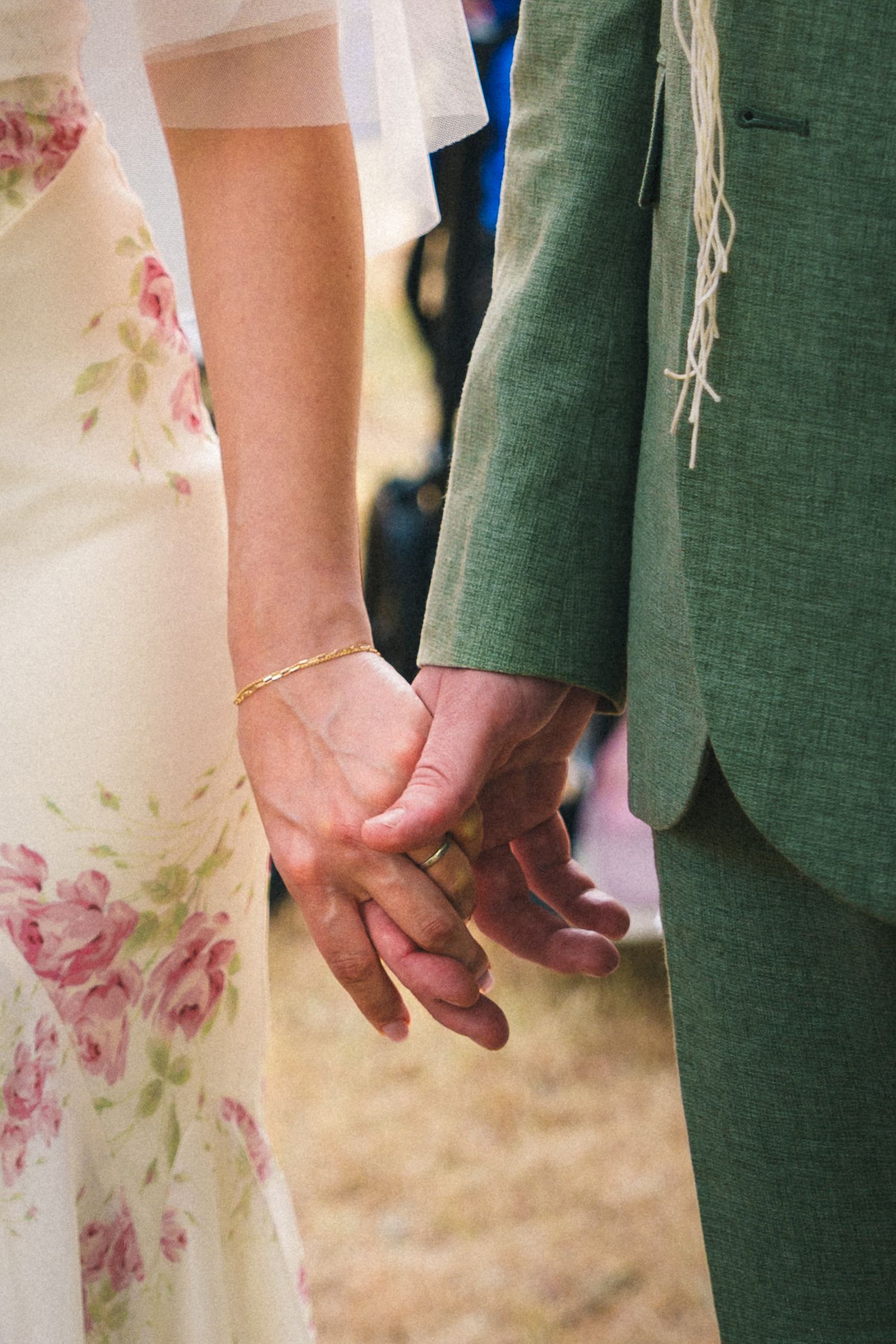 Bride and groom holding hands, wearing floral dress and green suit at an outdoor wedding ceremony.