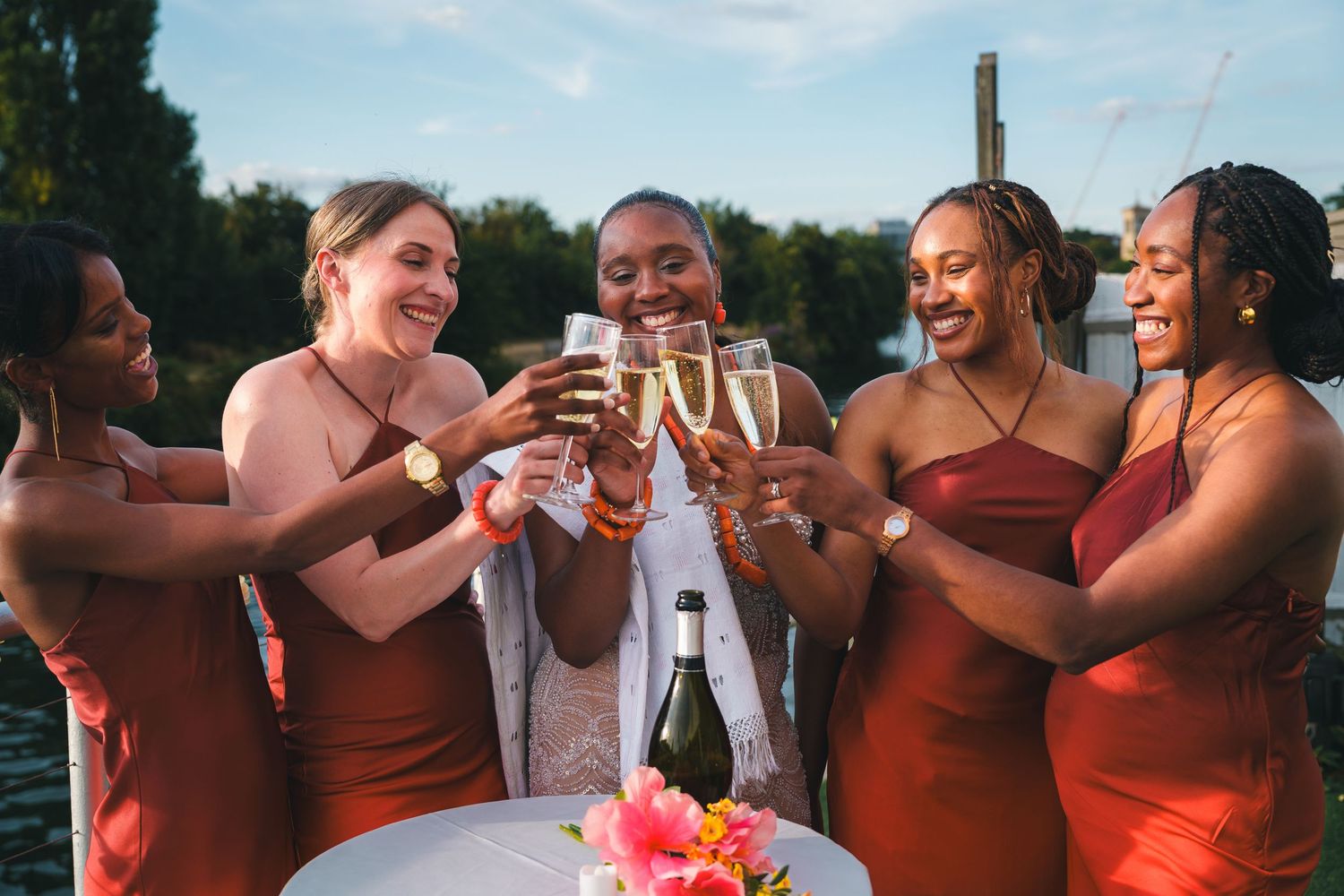 Five women in rust-colored dresses toasting with champagne glasses outdoors near water at a celebration.