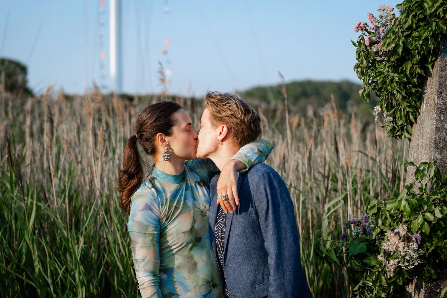Two people sharing a romantic kiss outdoors among tall green reeds and wildflowers in soft evening light.