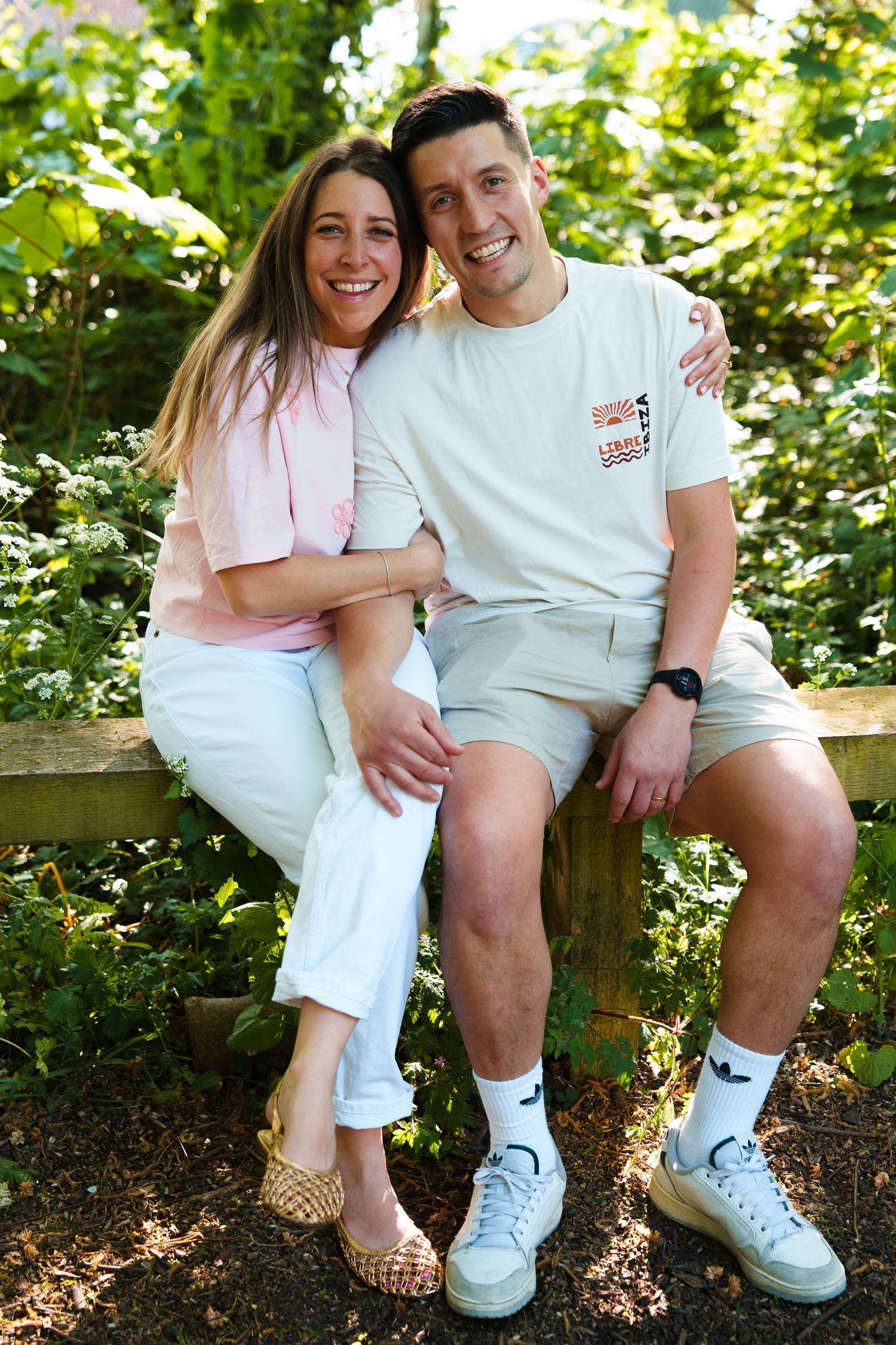 A smiling couple sits together on a wooden bench surrounded by lush green foliage in a garden setting.