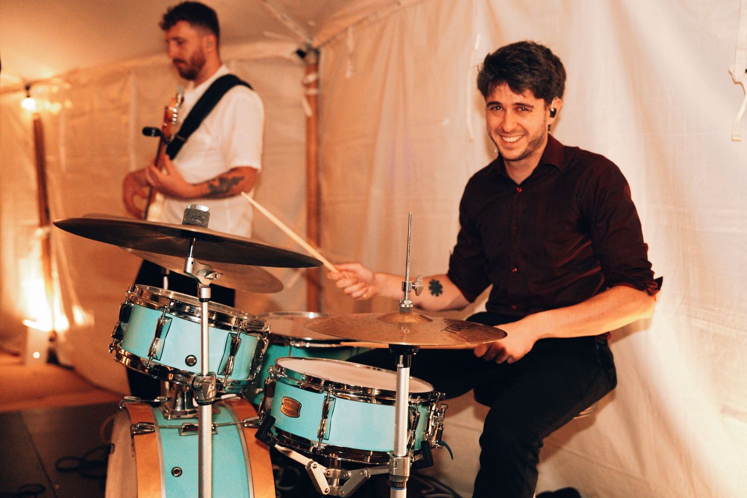 Smiling drummer playing light blue drum kit on stage with guitarist performing in background.