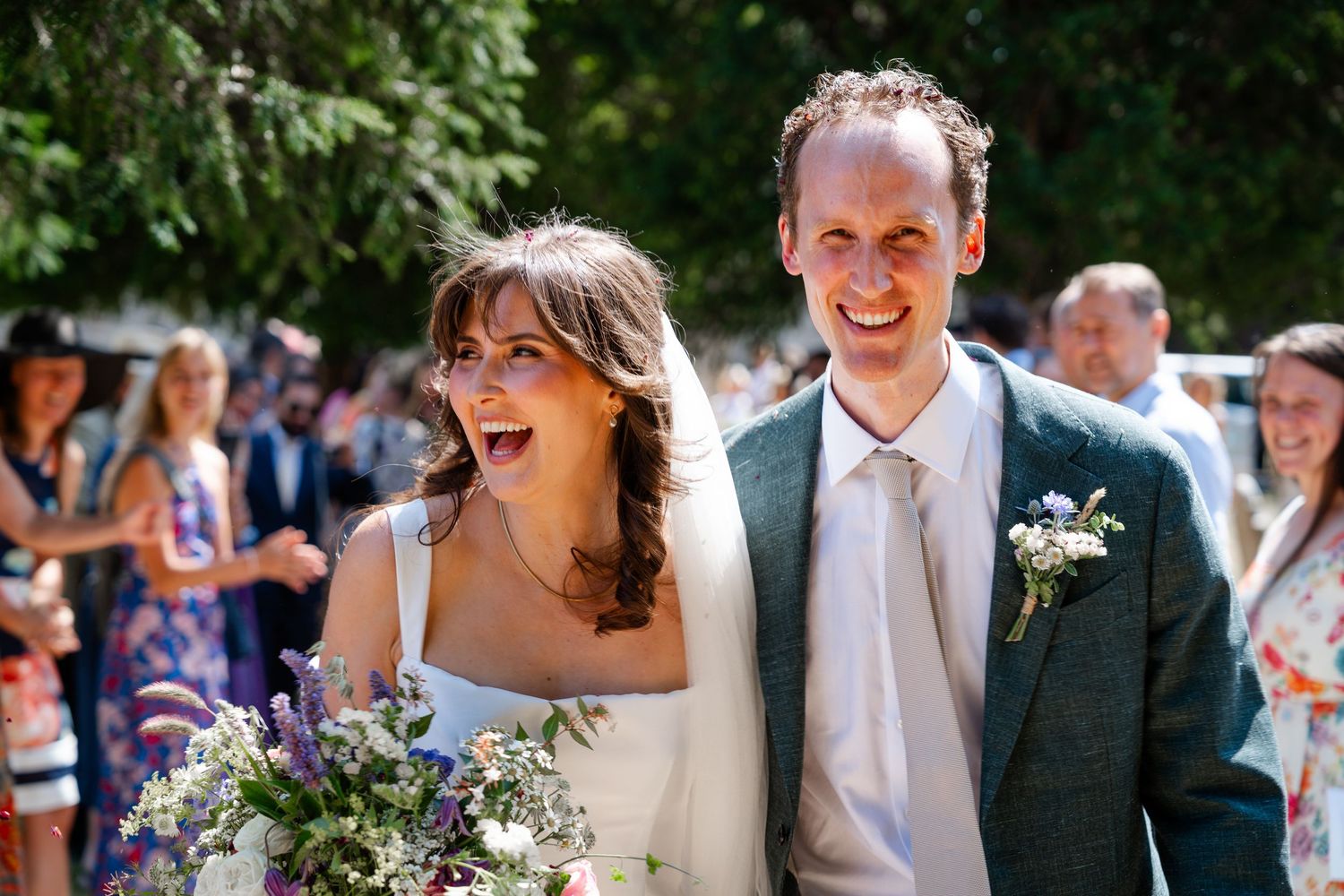 Happy bride and groom walking together after wedding ceremony, bride holding wildflower bouquet, guests celebrating in background.