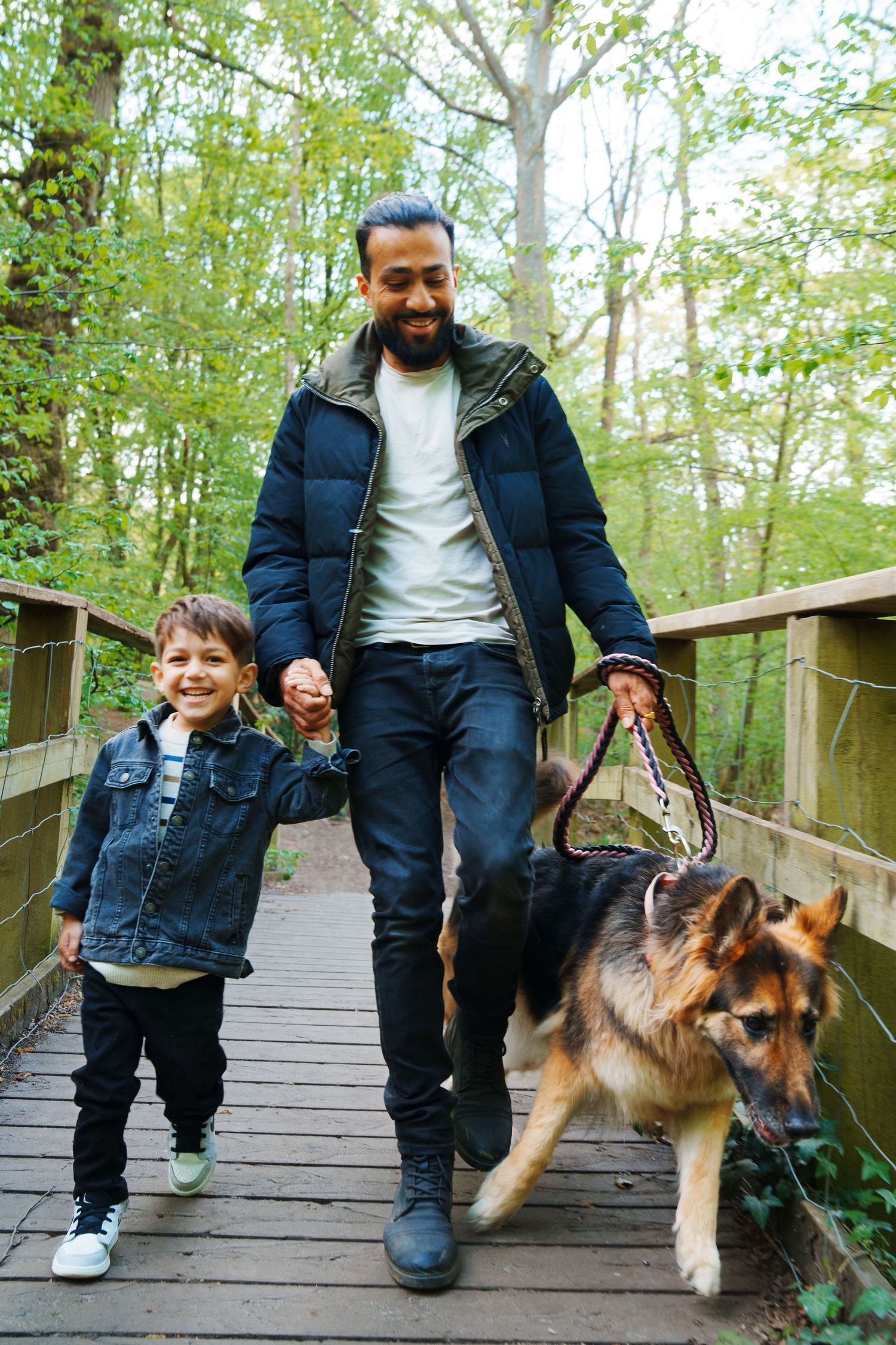 A smiling man and young boy walk a German Shepherd dog on a wooden bridge path through a lush green forest.