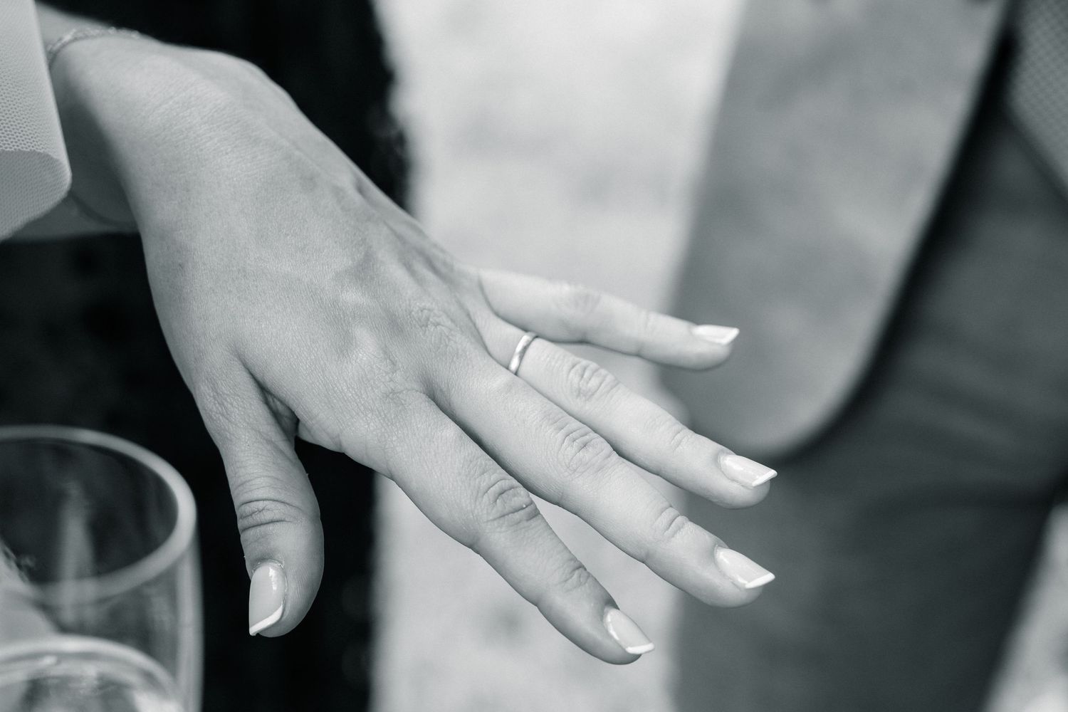 Close-up black and white photo of a bride's hand displaying a delicate wedding band with French manicured nails.