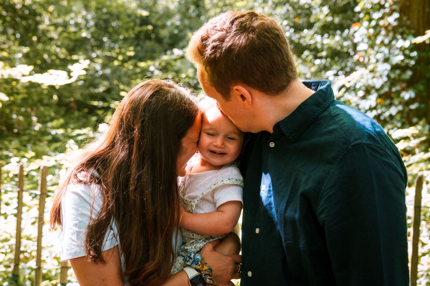 Happy family moment with parents kissing smiling baby outdoors in lush green garden setting.