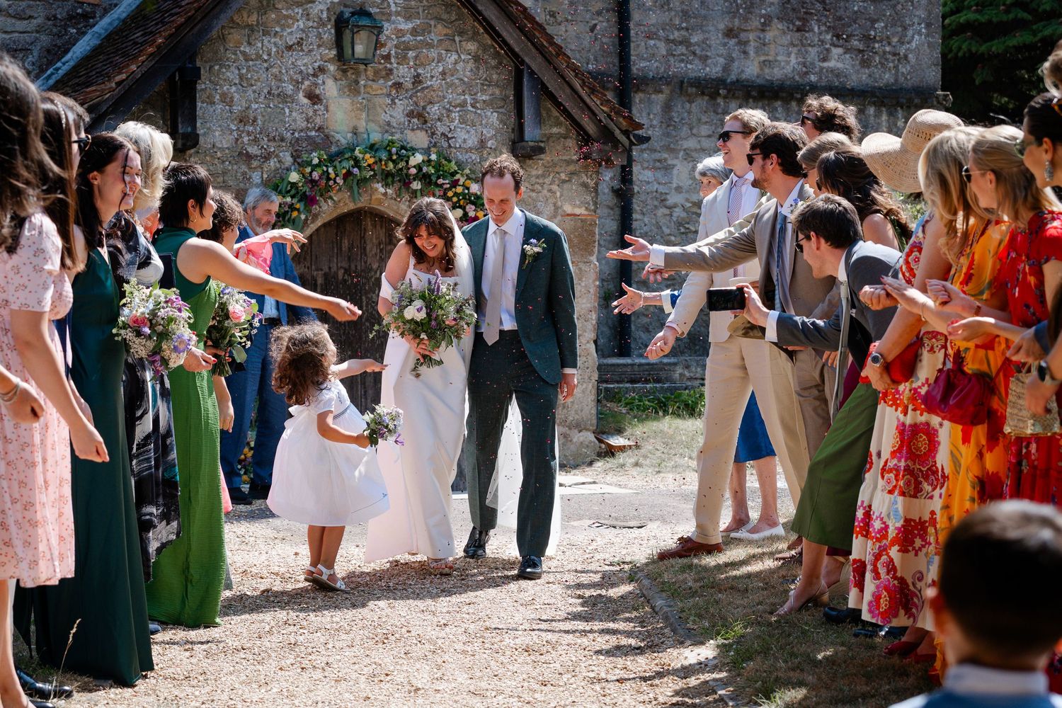 Bride and groom walk through cheering guests outside a stone church, with a flower girl and bridesmaids holding bouquets.