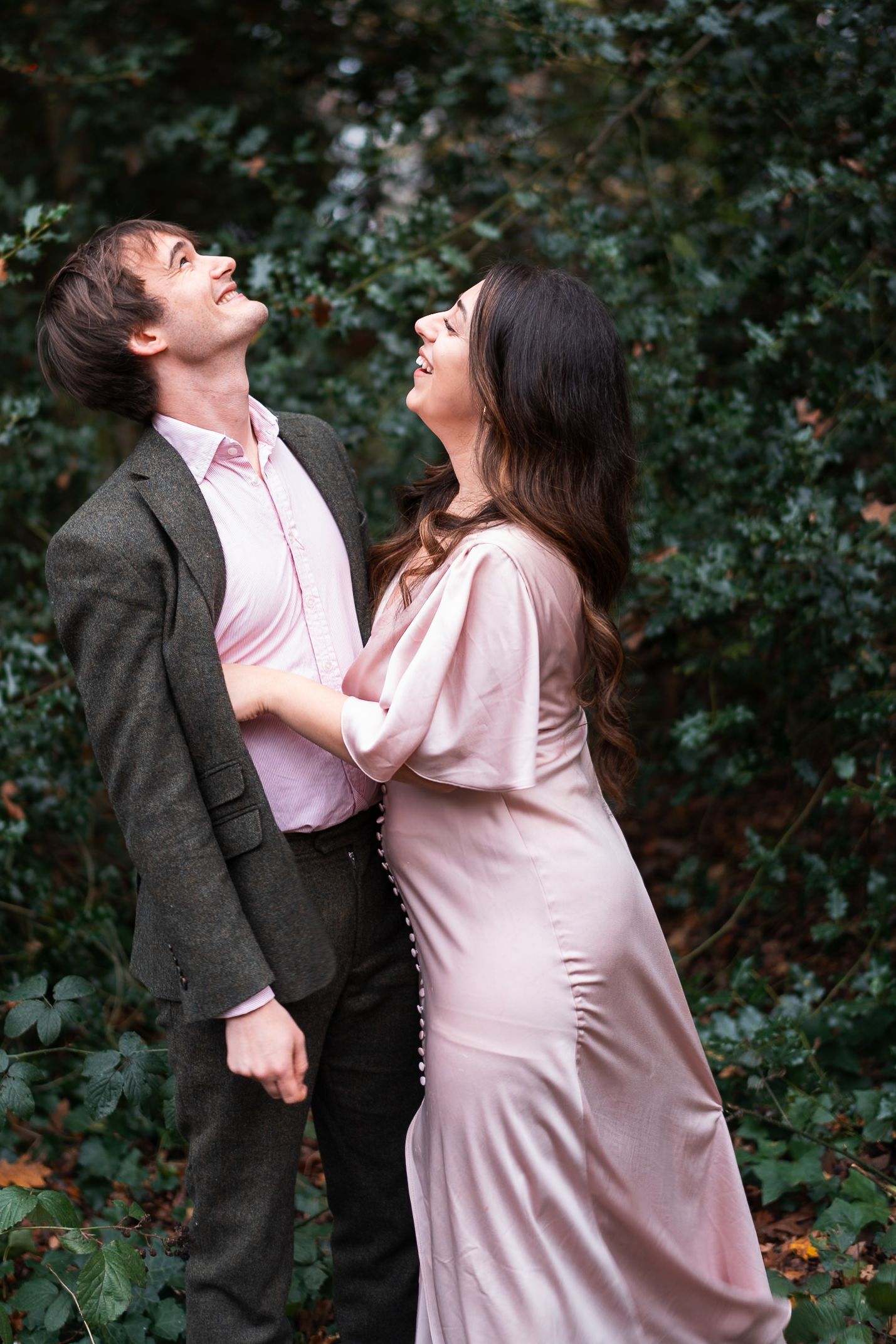 Couple in a romantic outdoor photoshoot among green foliage wearing pink and grey attire.