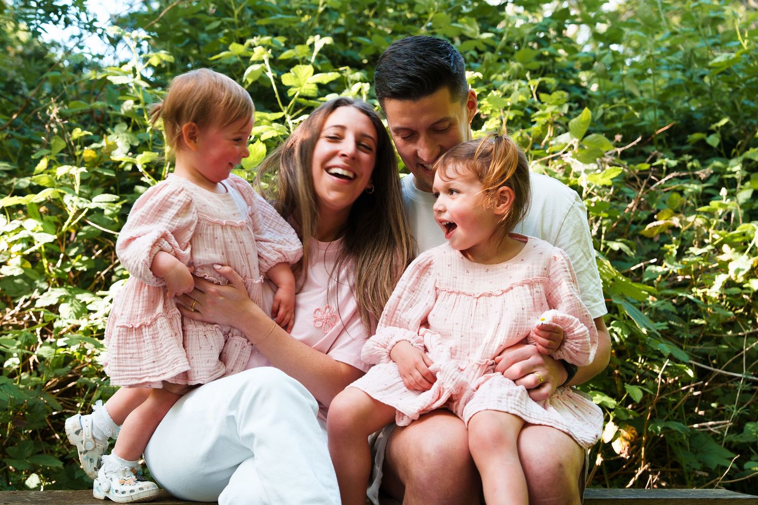 Happy family with two young girls in pink dresses laughing together outdoors surrounded by lush green foliage.