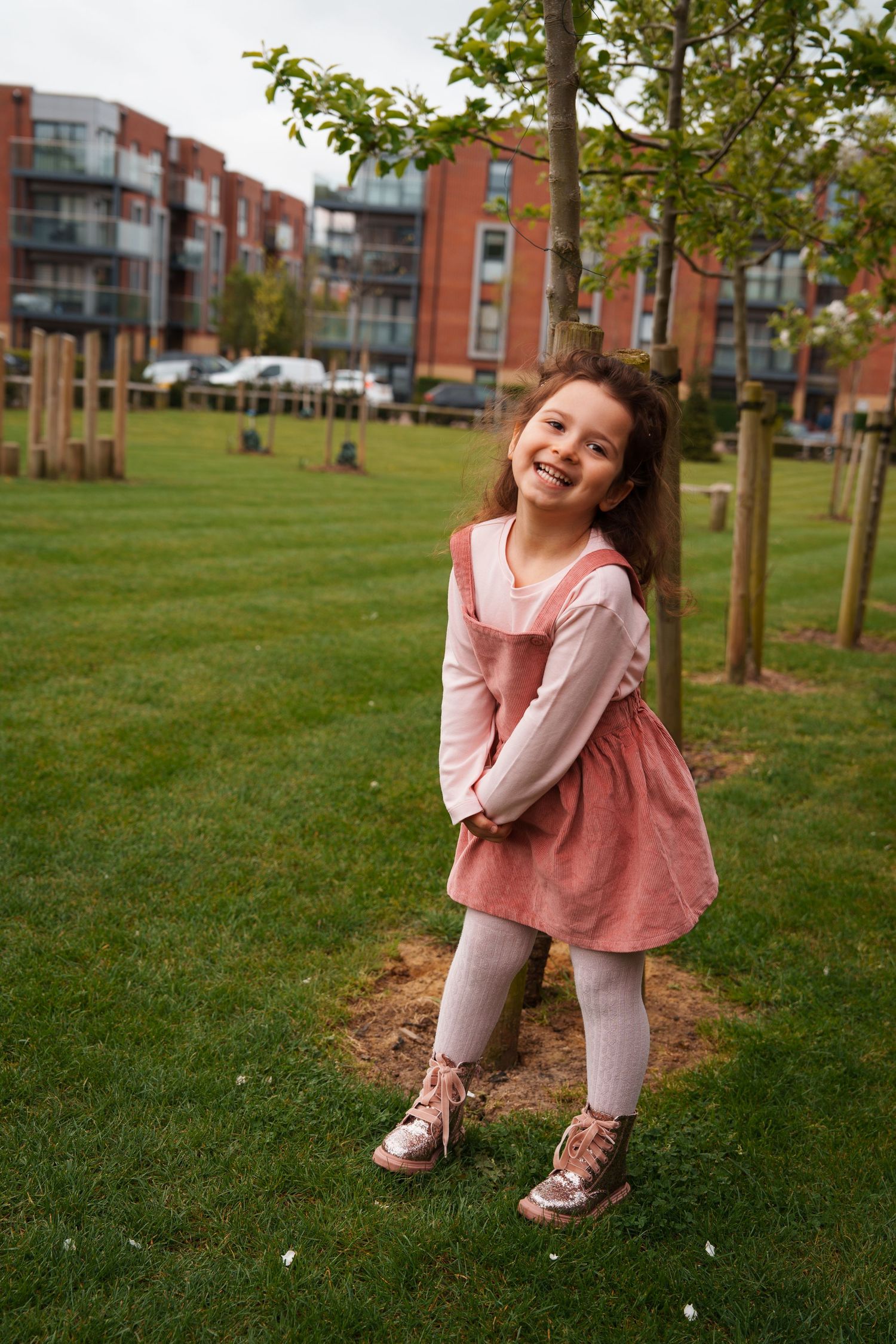 Smiling young girl in pink dress and metallic boots posing by a tree in a urban park setting.