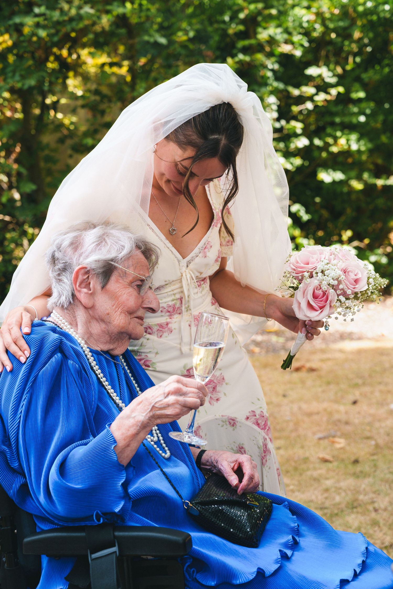 Bride in white veil leans over elderly woman in blue dress holding champagne glass at outdoor wedding ceremony.