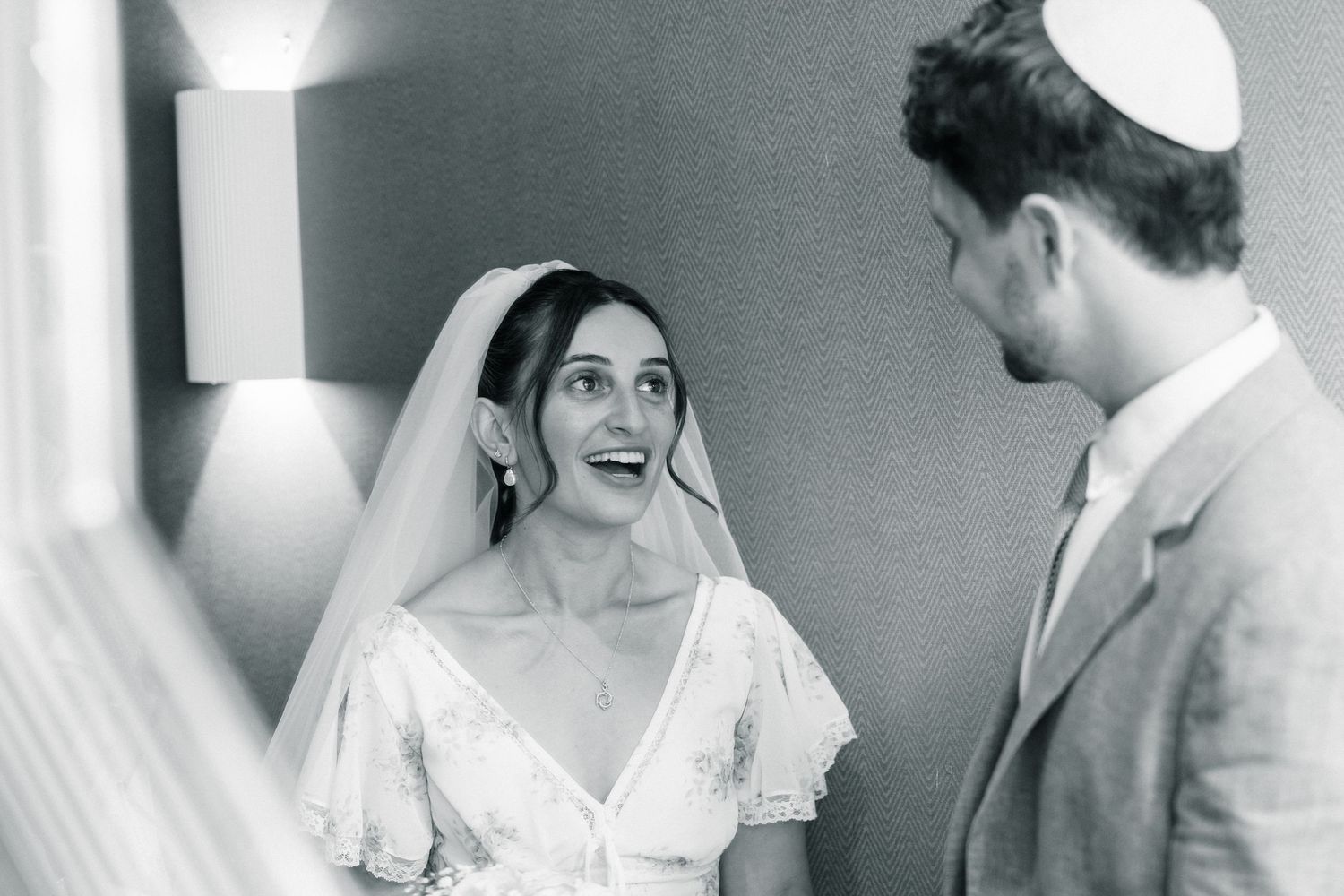 Black and white photo of a smiling bride in veil and groom in kippah sharing a joyful moment at their Jewish wedding.