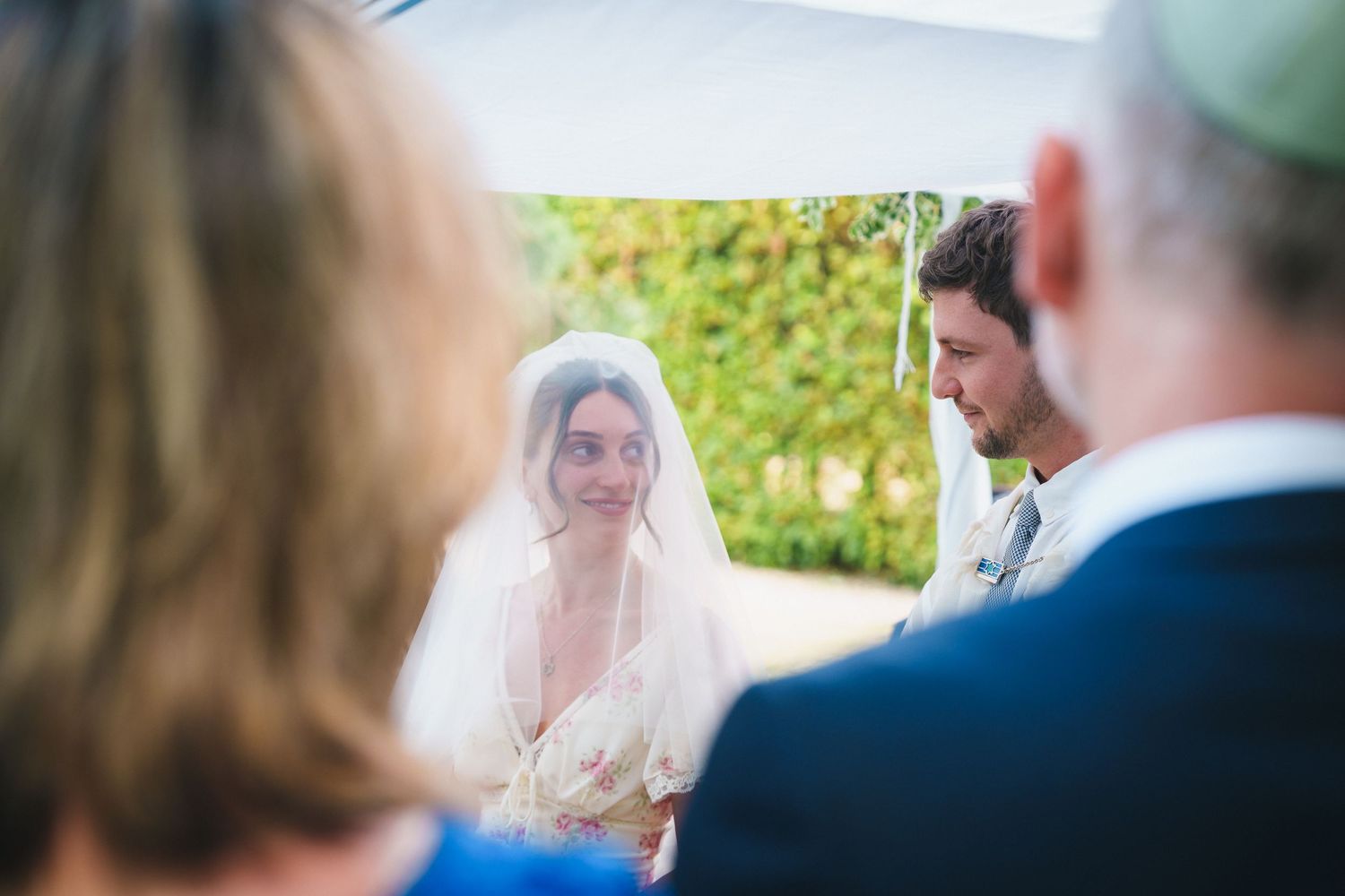 Bride in veil and groom share an intimate moment during an outdoor Jewish wedding ceremony under a chuppah.