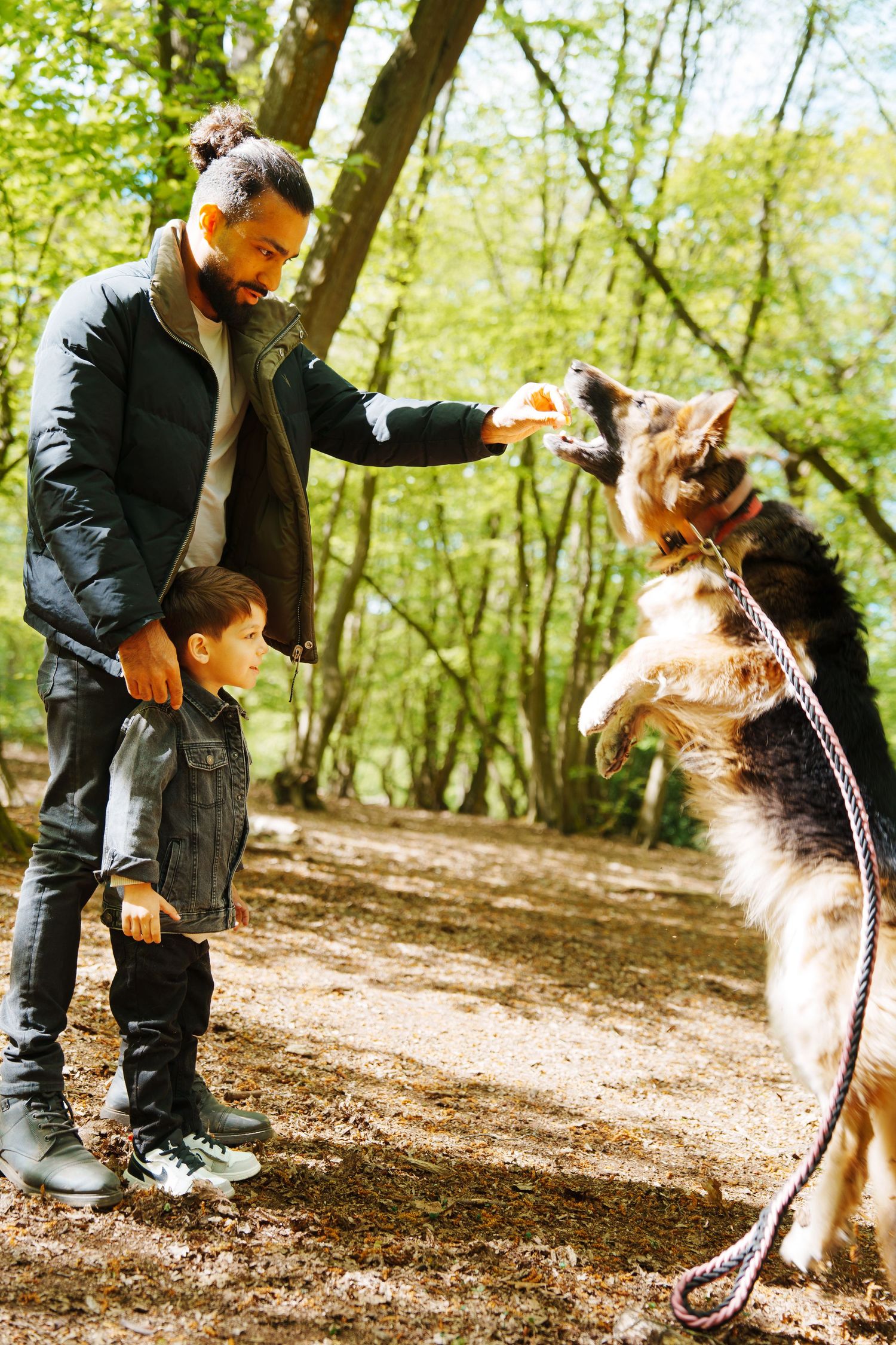 Man and child feeding a jumping German Shepherd dog on a leash in a lush green forest park.
