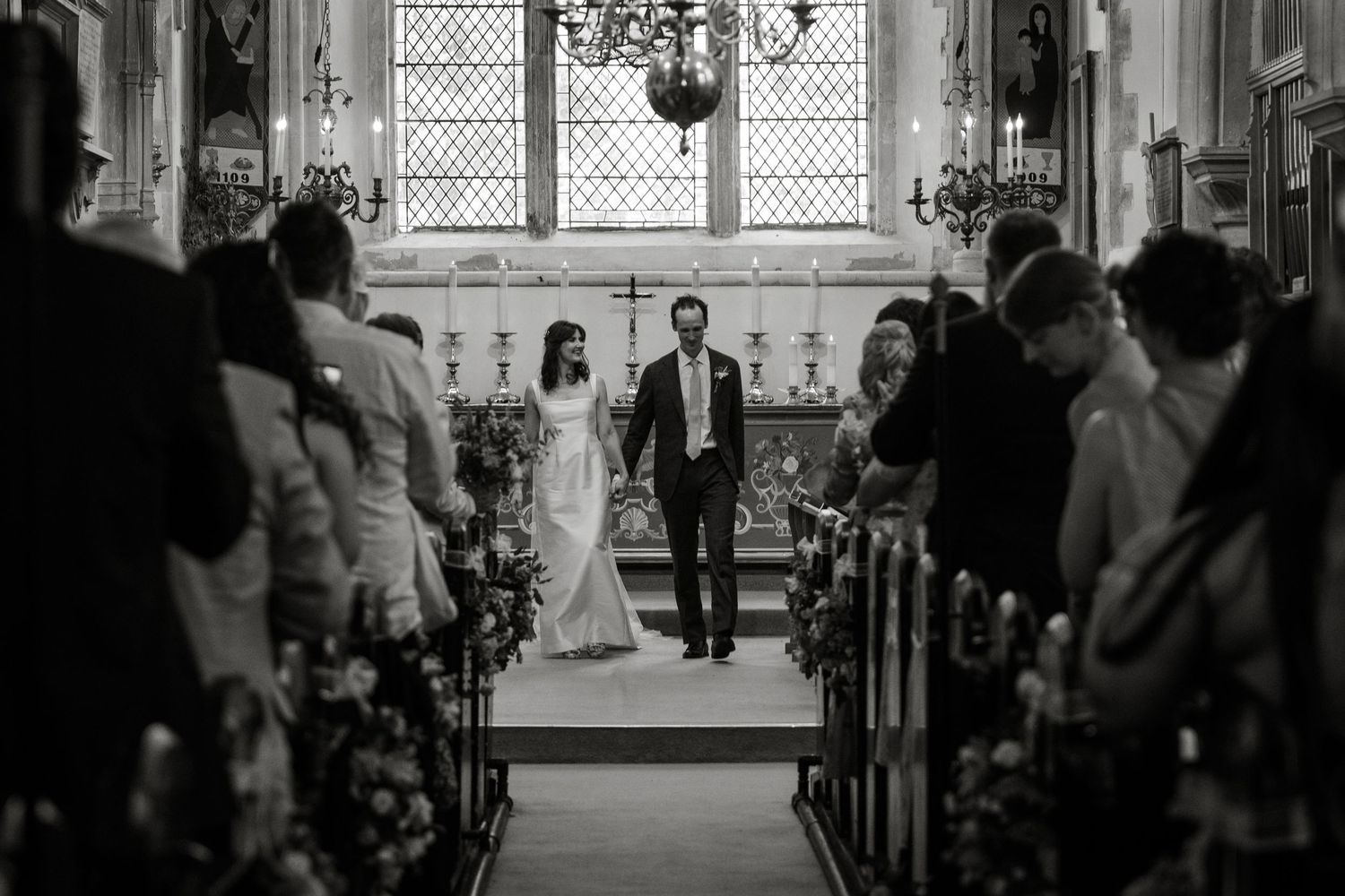 Black and white wedding photo of bride and groom walking down church aisle during ceremony.