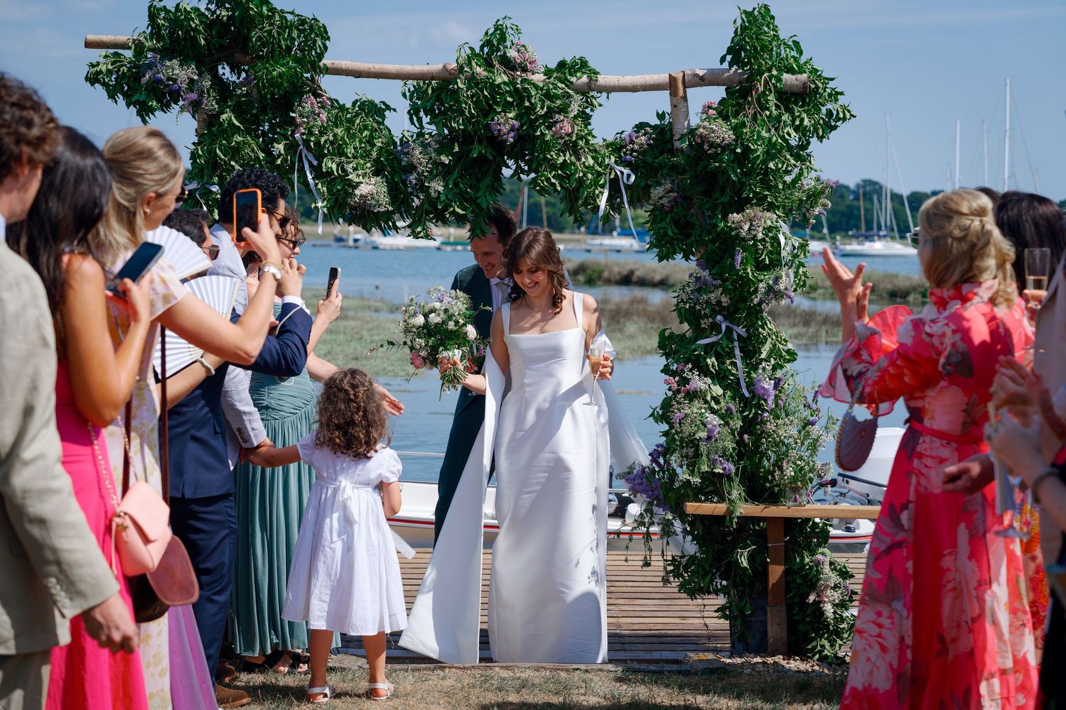 Bride and groom walk down outdoor aisle under floral arch by waterfront, surrounded by cheering wedding guests.