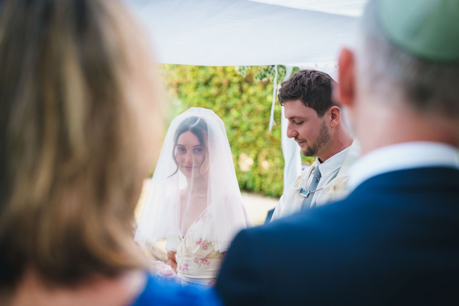 Bride and groom stand under wedding chuppah during outdoor Jewish wedding ceremony.