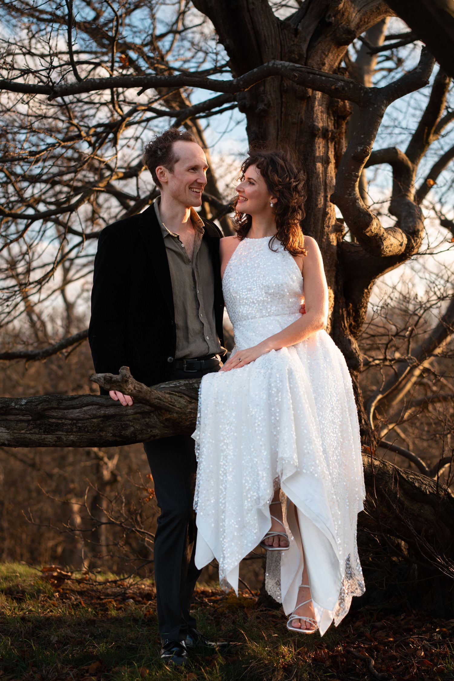 A white dress sparkles in the evening light as two people share a moment under a tree.