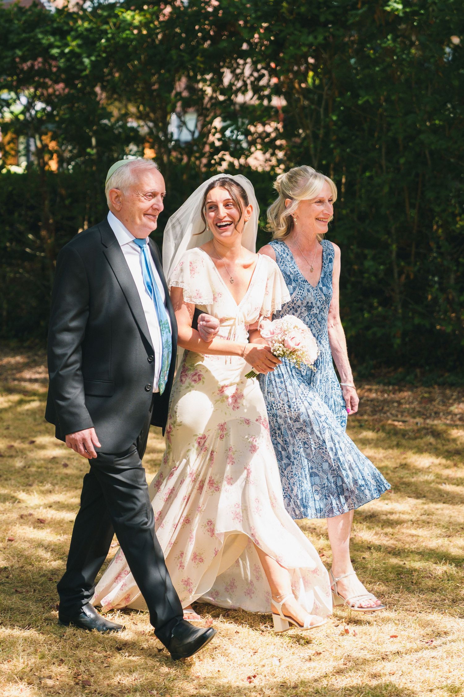 Bride in floral wedding dress walks with parents through sunny garden, laughing joyfully while holding pink bouquet.