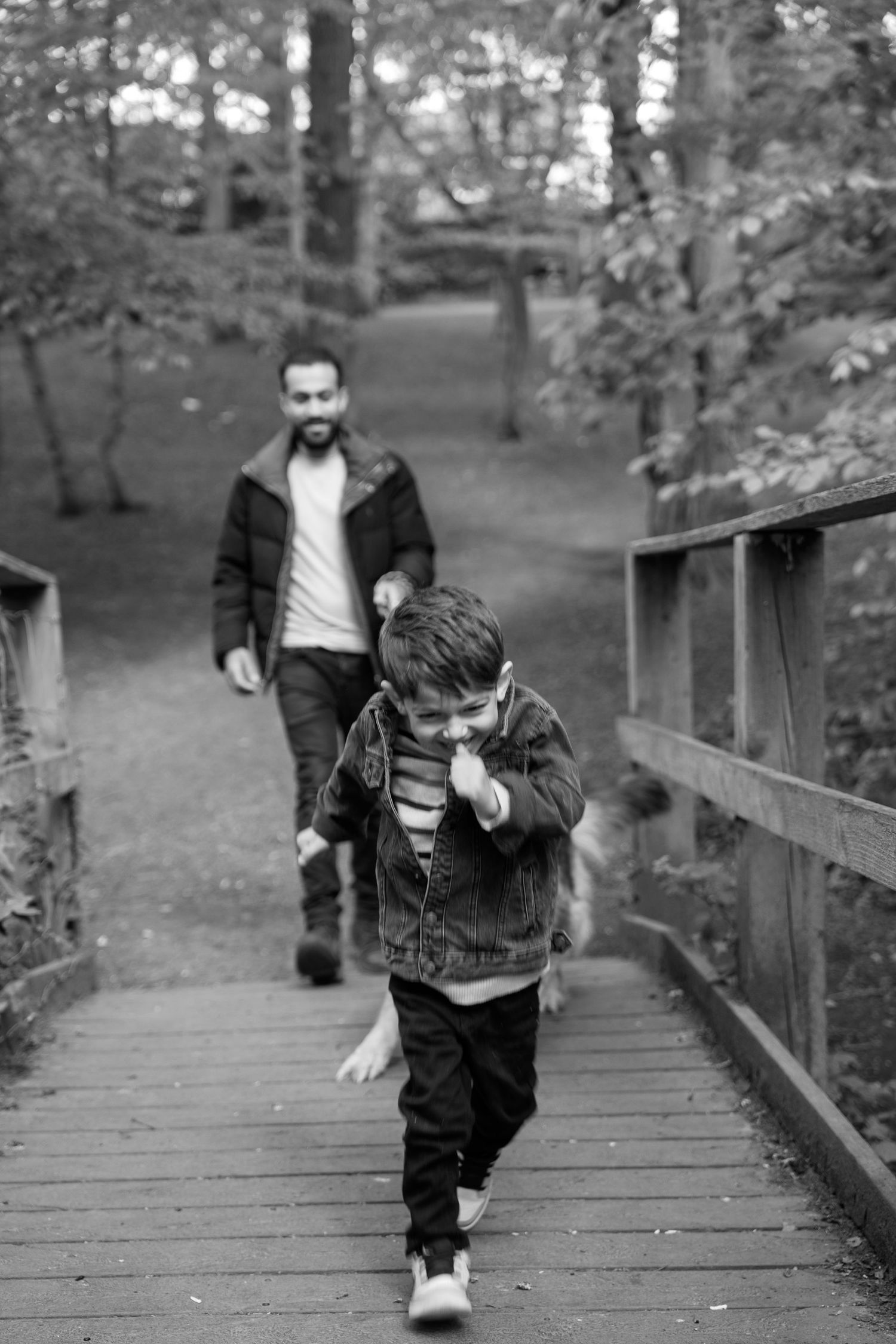 Black and white photo of a young boy running on a wooden bridge path in a park with a smiling man walking behind him.
