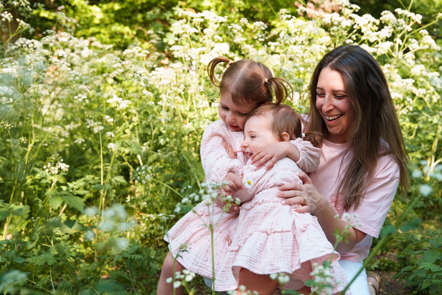 A smiling mother holds her baby and young daughter in a lush garden filled with white wildflowers.