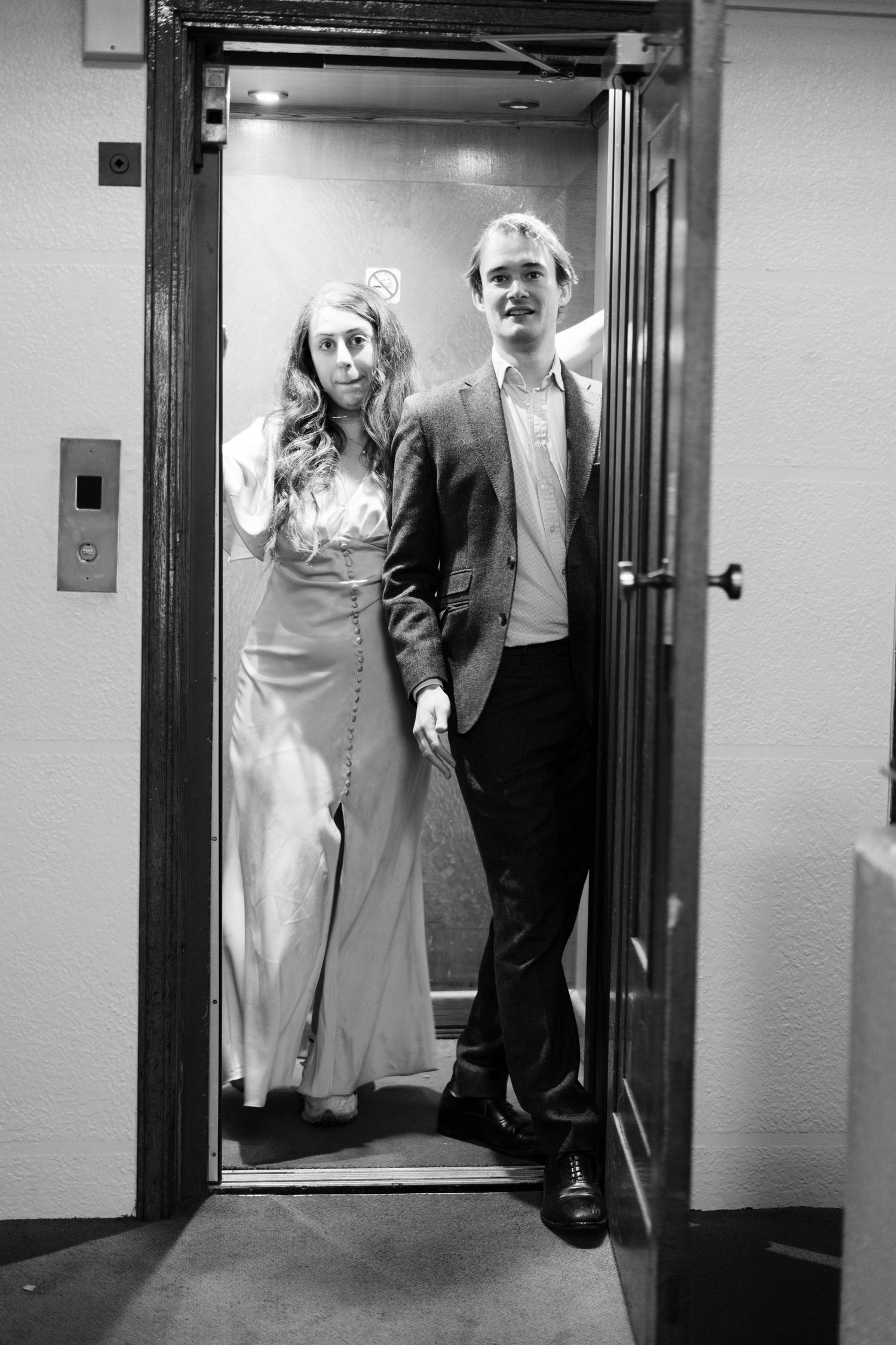 A black and white portrait of a couple in formal attire standing in an elevator doorway.