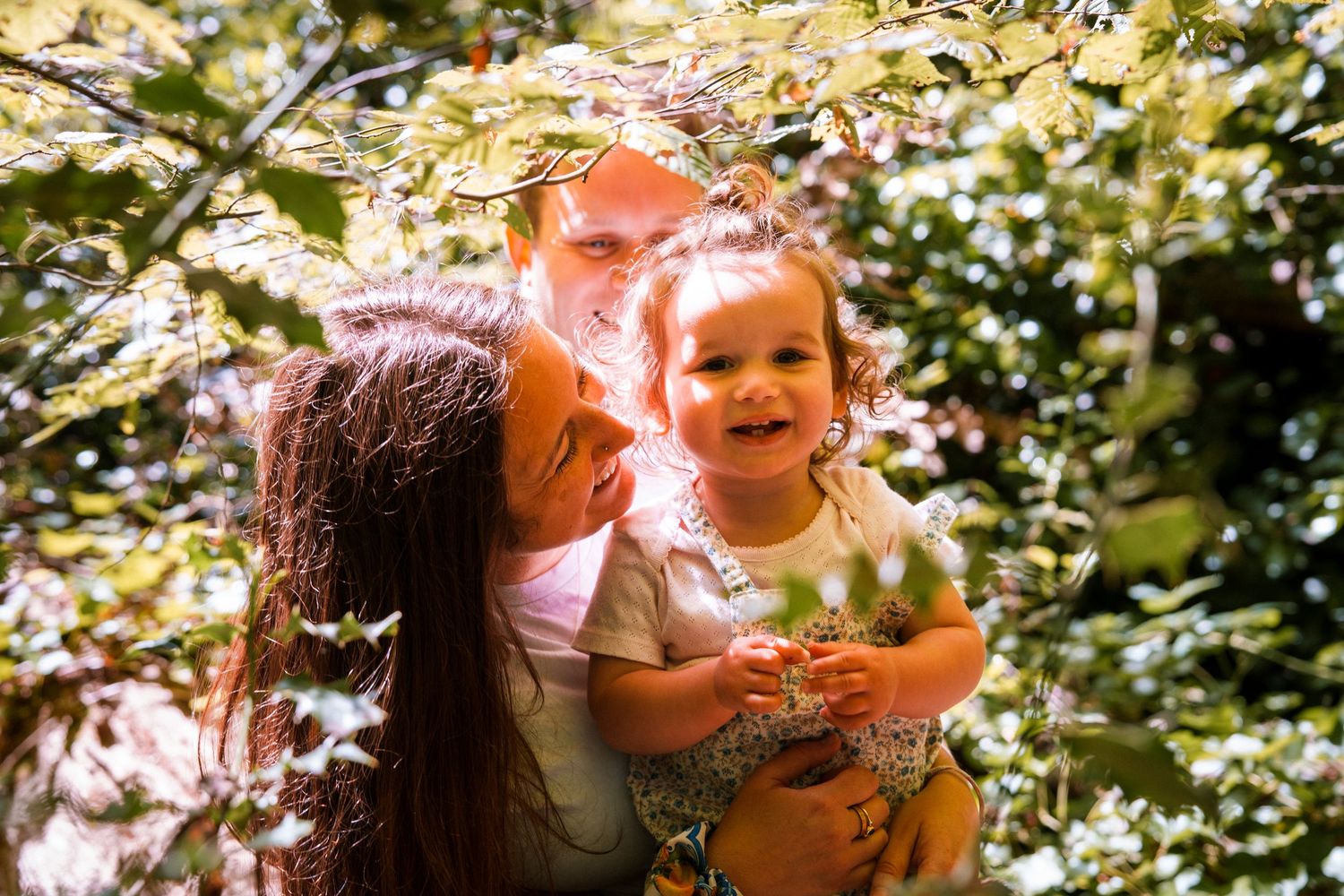 Happy family with parents and toddler girl smiling among green leaves in a sunlit garden.