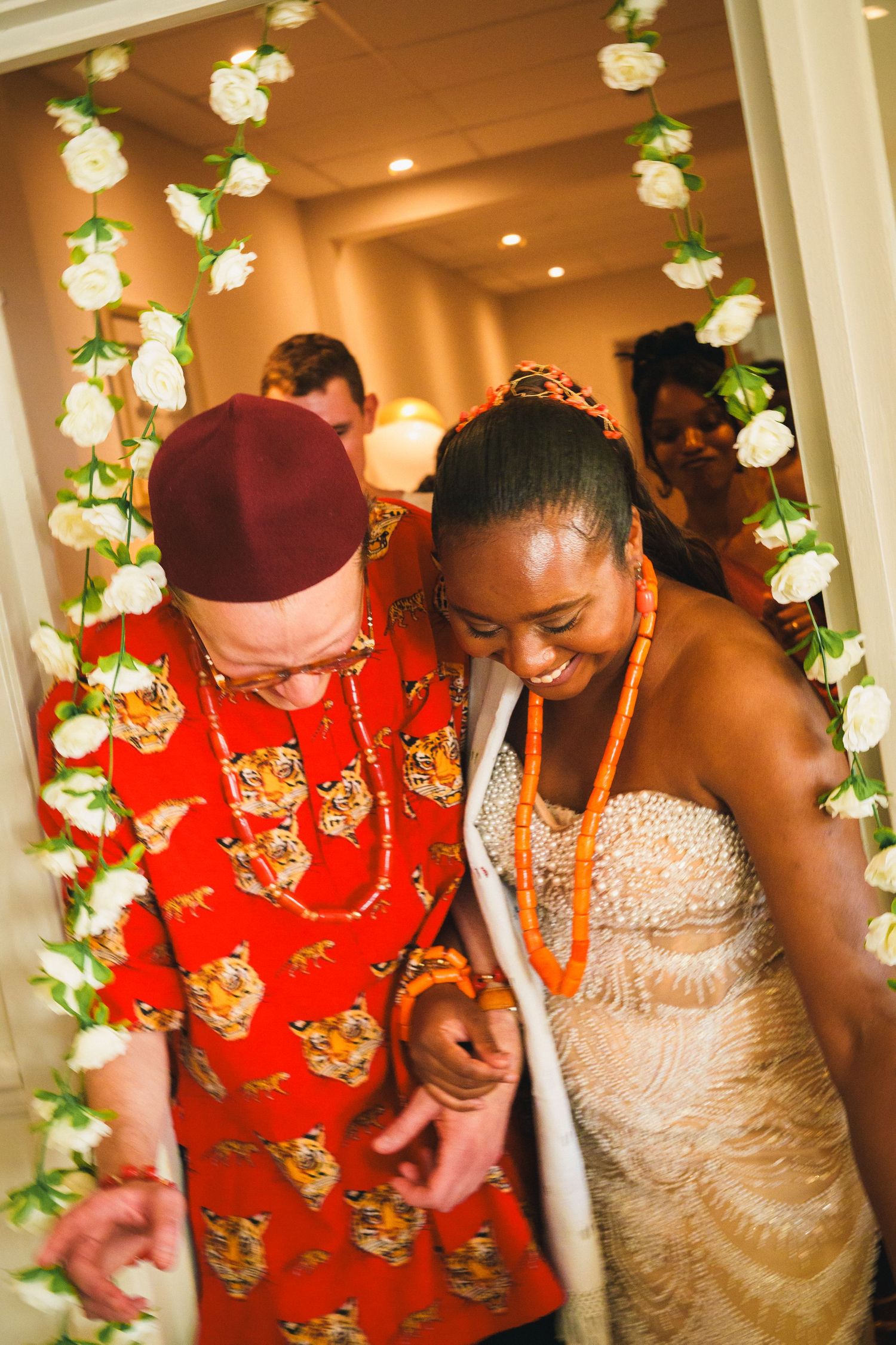 Couple in Nigerian traditional wedding attire walking through floral arch, groom in red tiger print top and bride in beaded gown.