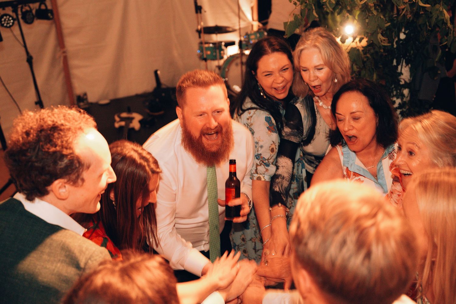 A joyful group of people laughing and celebrating together at an outdoor party, centered around a bearded man holding a beer.