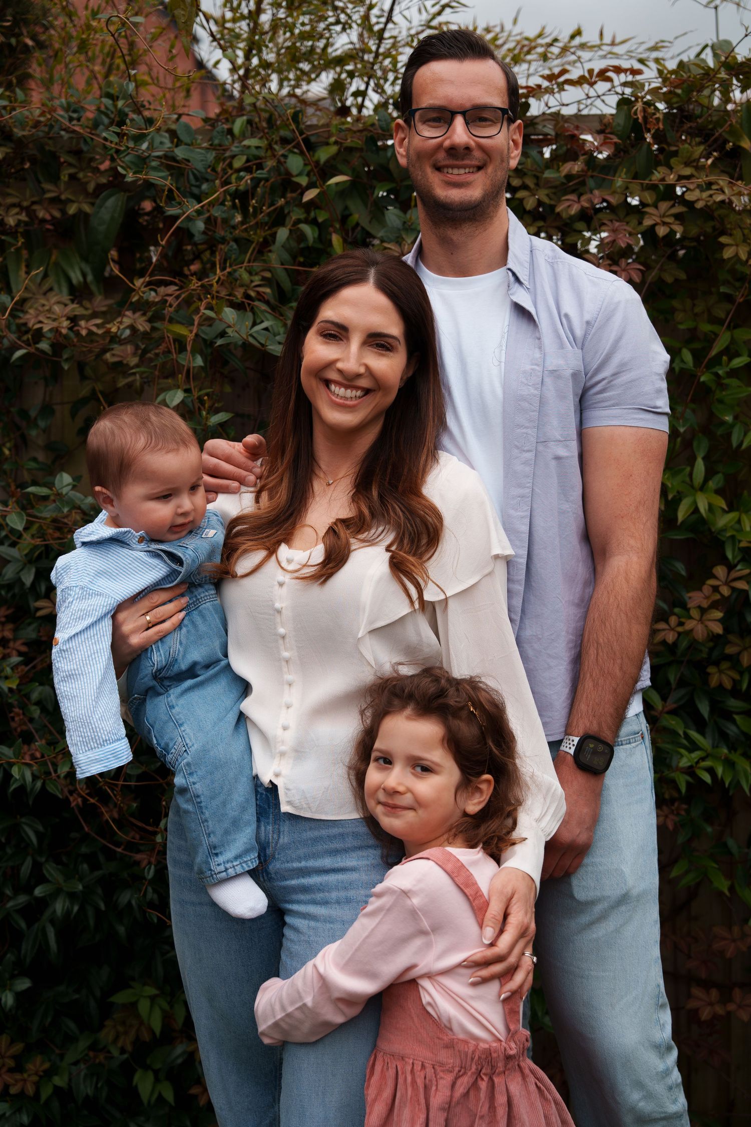 Happy family of four posing outdoors, parents with baby and young girl in pink dress, surrounded by green foliage.