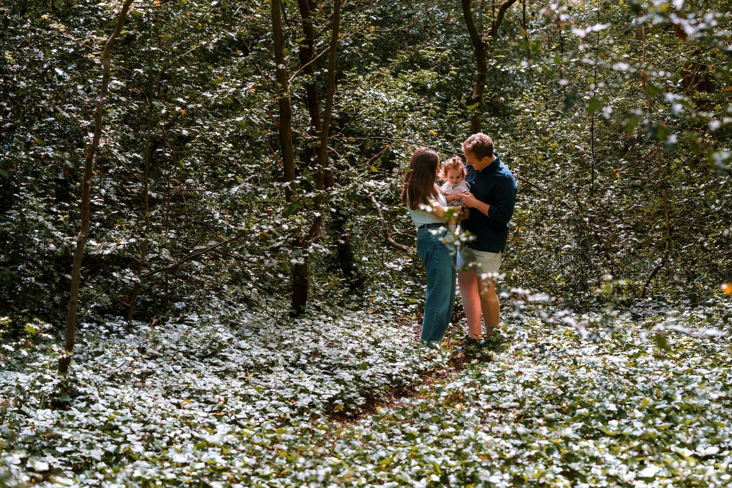 Couple embracing in a sunlit forest clearing covered with white wildflowers surrounded by lush green trees.