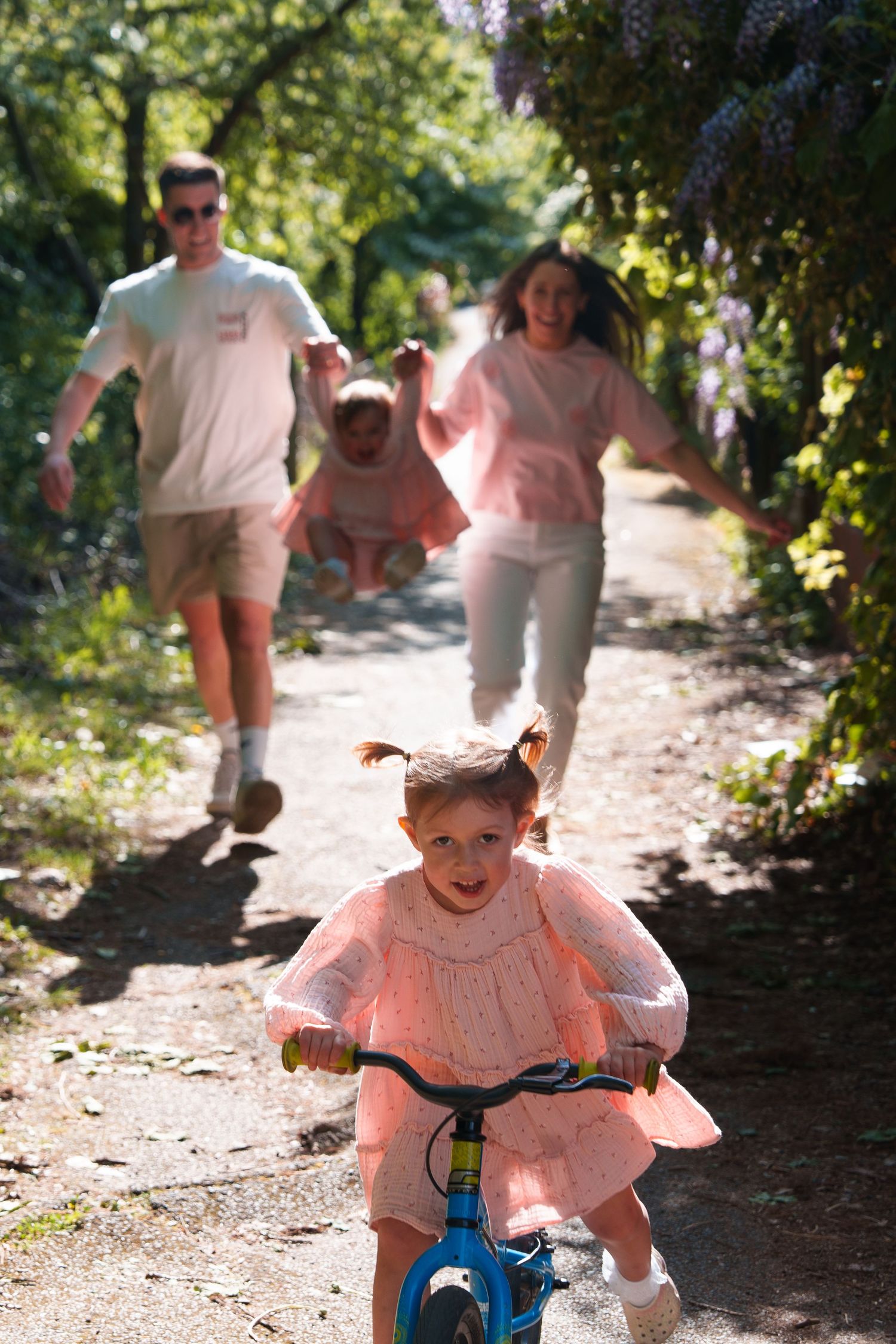 A young girl in a pink dress rides a bike on a garden path while her family runs behind her on a sunny day.