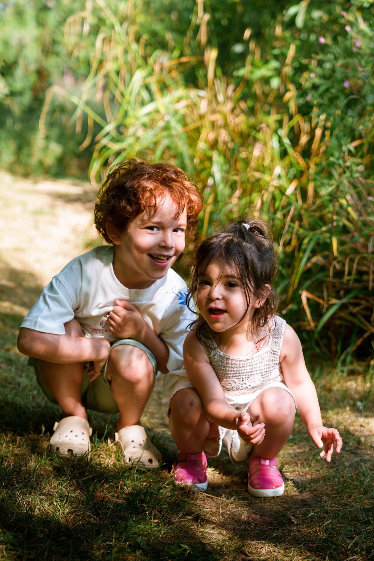 Two young children squat together outdoors in a sunny garden, a red-haired boy and a toddler girl smiling joyfully.