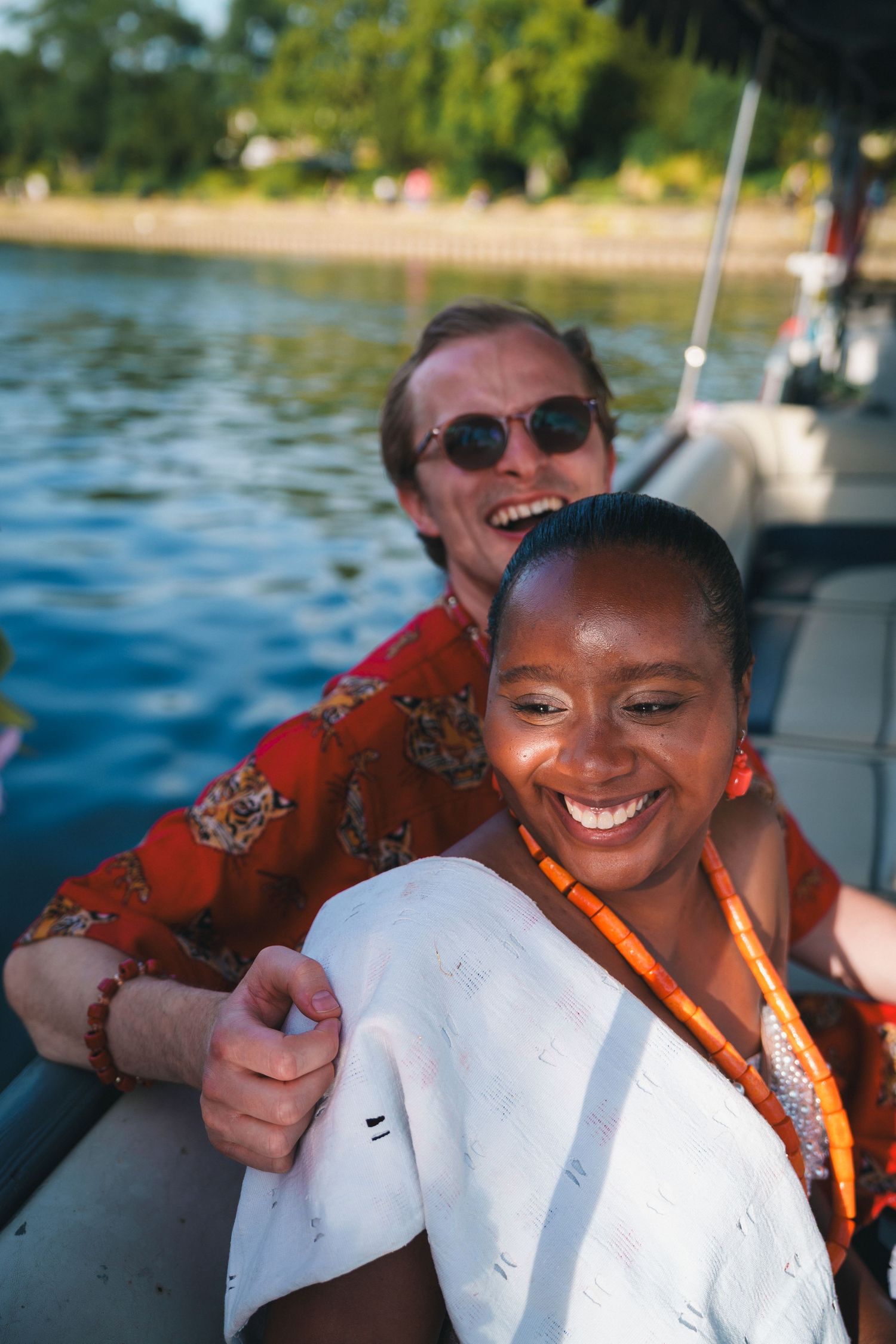 A smiling interracial couple enjoying a sunny boat ride near a green shoreline, woman in white top with orange beaded necklace.