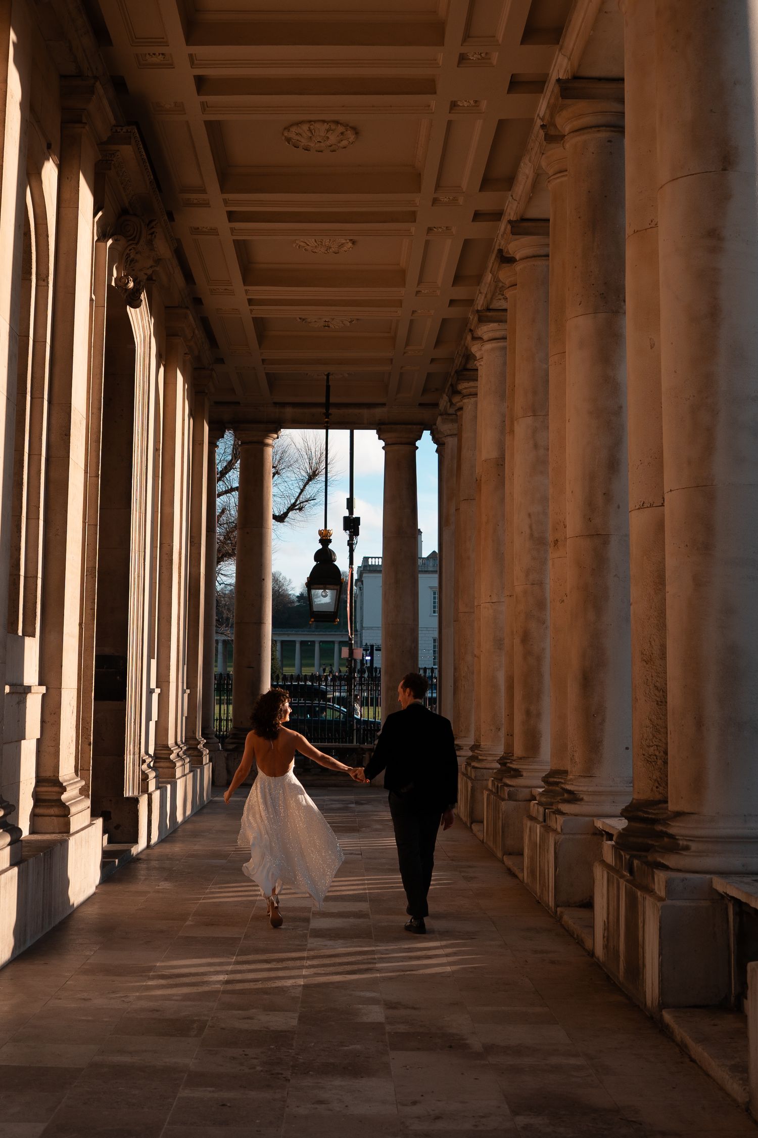Long architectural colonnade with classical columns and figures walking in warm sunlight.