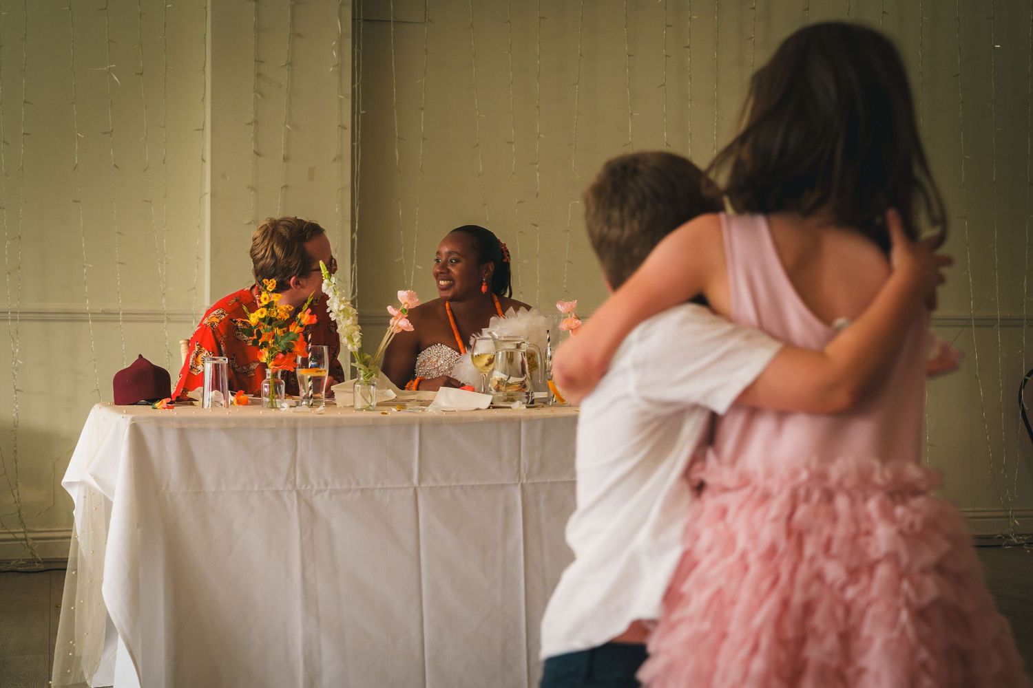 Two children embrace at a wedding reception while guests sit at a decorated head table in the background.