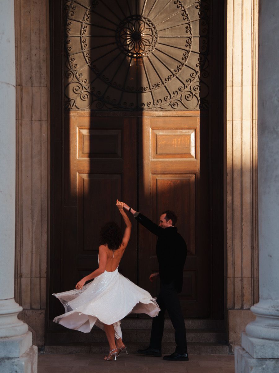 A couple dances elegantly before grand wooden doors, bride in flowing white backless gown, groom in black, golden light.