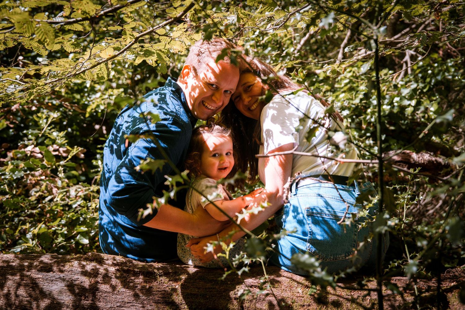 Family of three posing together among green tree branches in a sunlit forest setting.