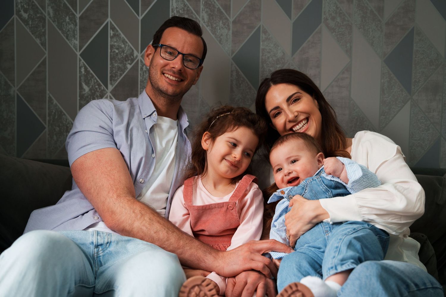 Happy family of four sitting together on couch, parents with two young children smiling at camera in casual clothing.