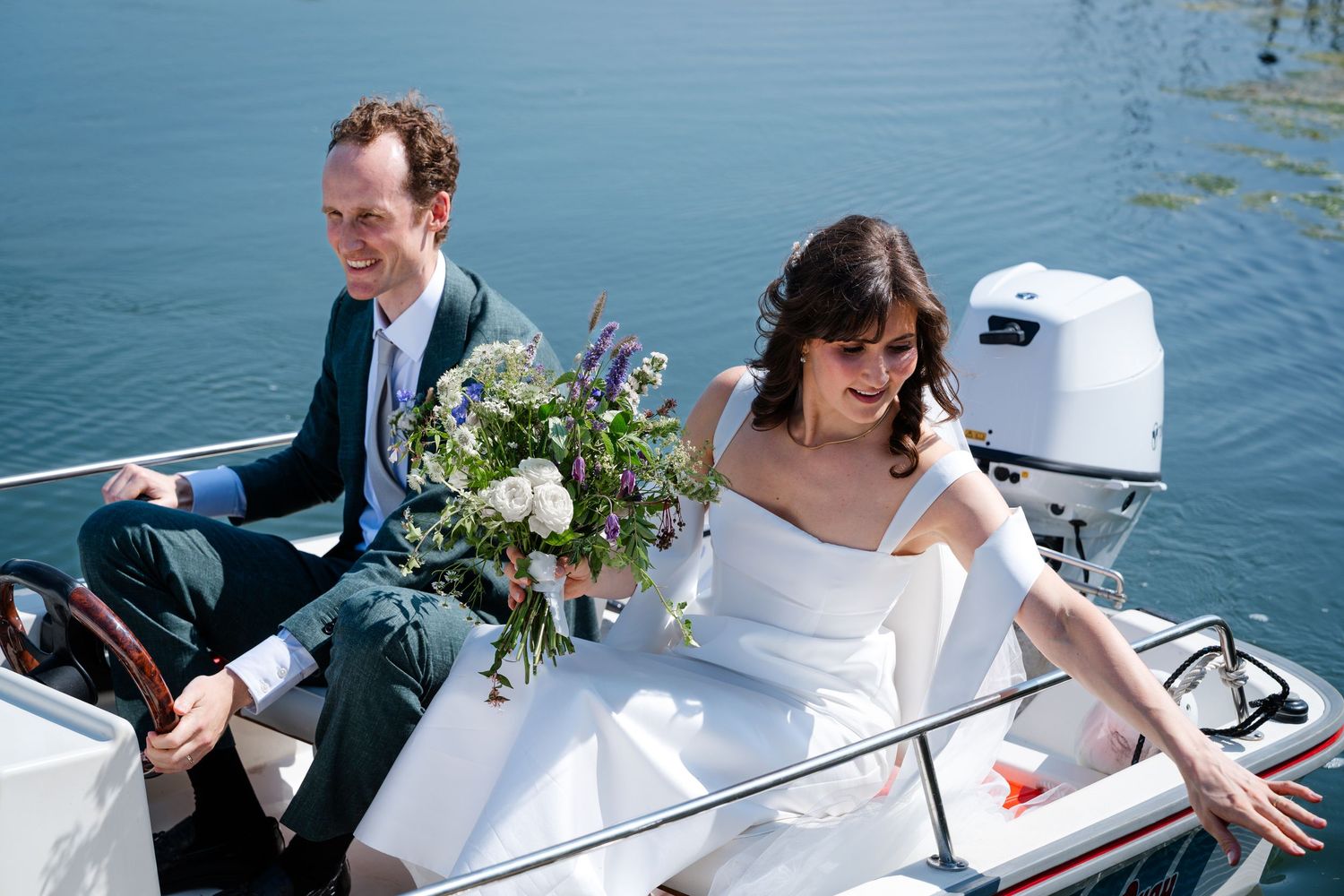 Bride and groom sitting on a boat on calm blue water, bride holding a wildflower bouquet in a white wedding dress.