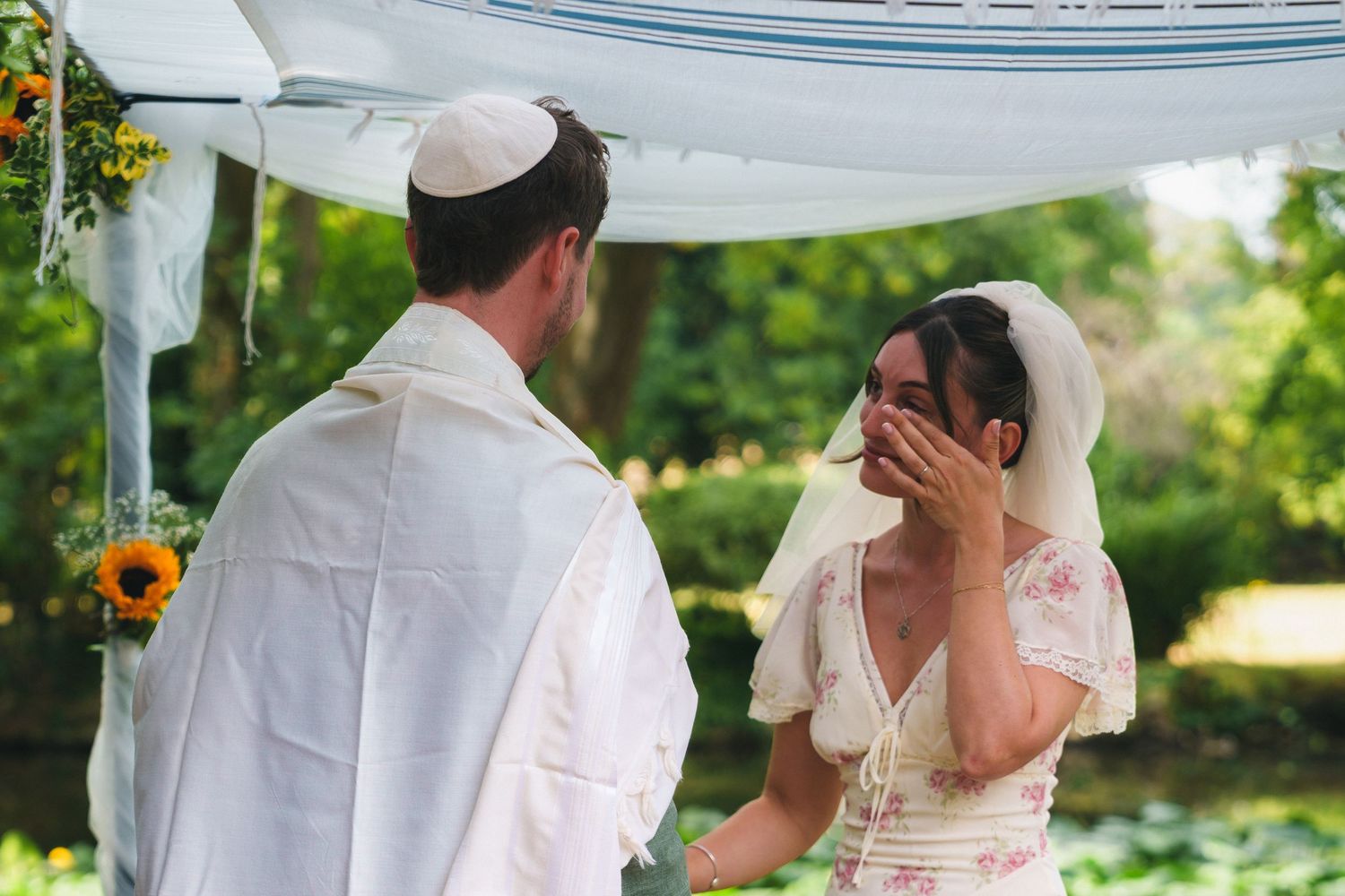 Jewish bride and groom sharing emotional moment under floral chuppah at outdoor wedding ceremony.