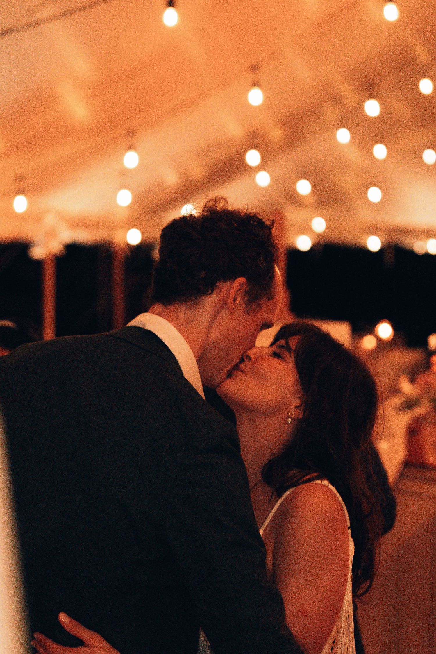 Couple sharing a romantic first dance kiss under warm string lights at an elegant evening wedding reception.