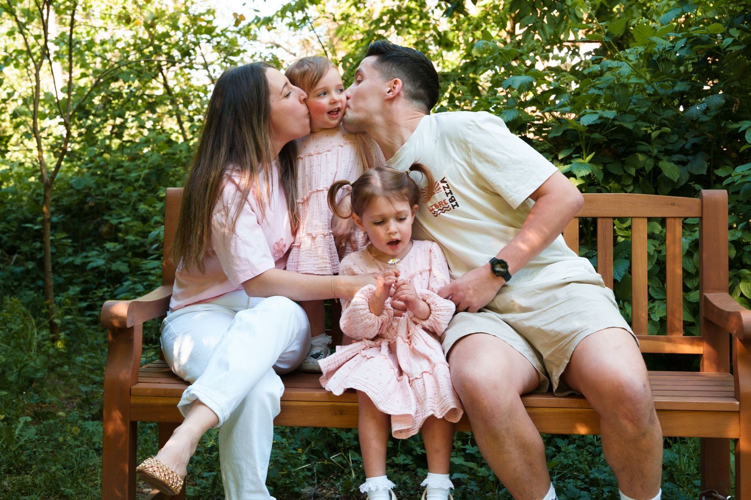 A happy family of four sits on a wooden bench outdoors, with parents kissing their two young daughters in pink dresses.