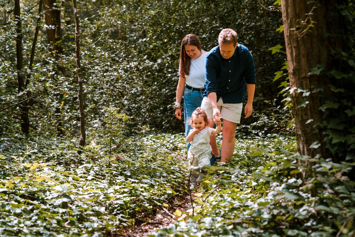 Family with young child hiking through lush green forest trail on a sunny day.