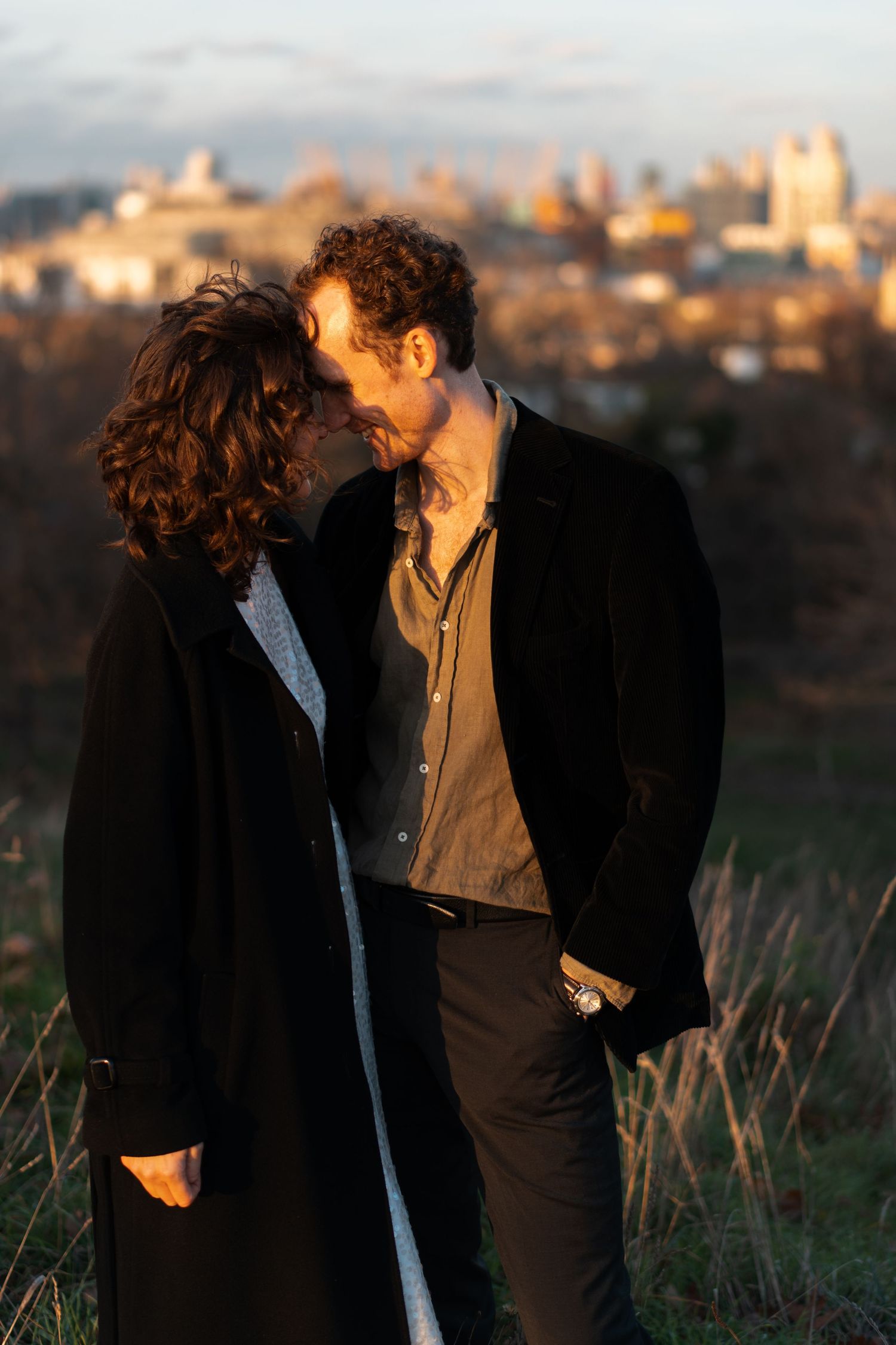 A romantic sunset photoshoot of a couple standing close together on a grassy hillside.