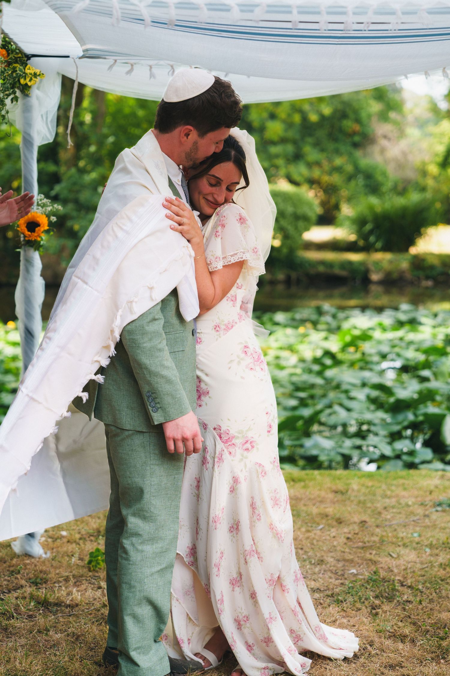 Groom in green suit embraces bride in floral dress under chuppah during outdoor Jewish wedding ceremony.