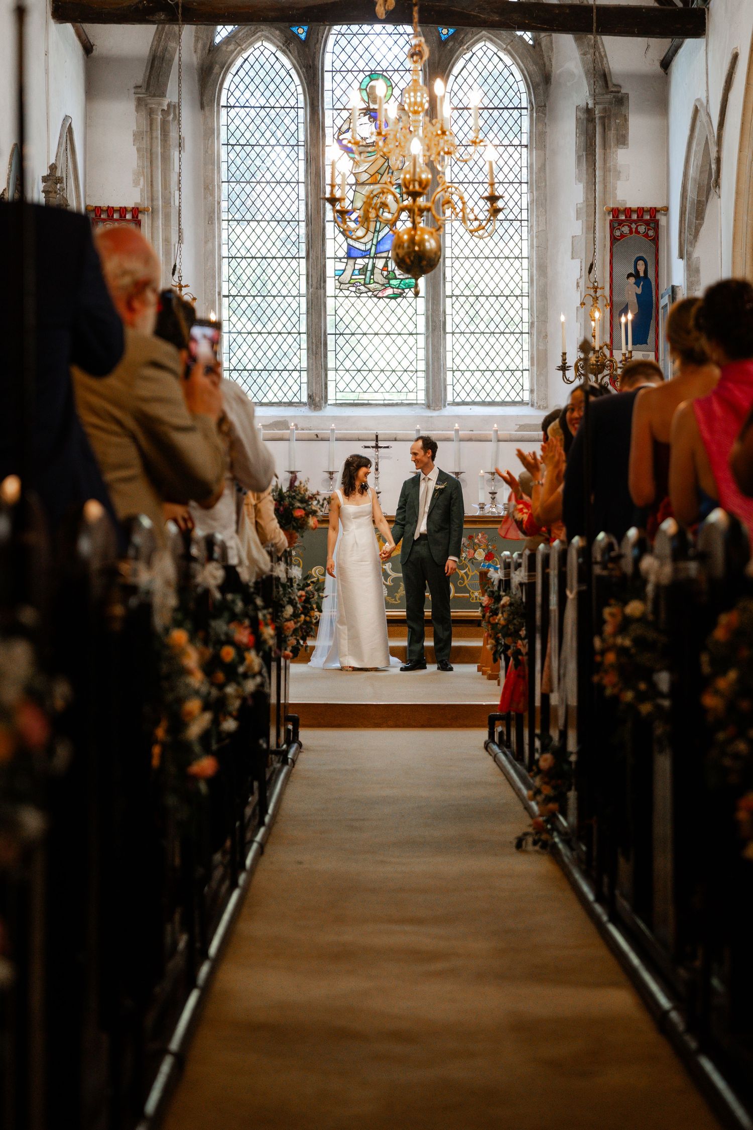 Bride and groom stand at church altar during wedding ceremony, flanked by flower-adorned pews and stained glass windows.