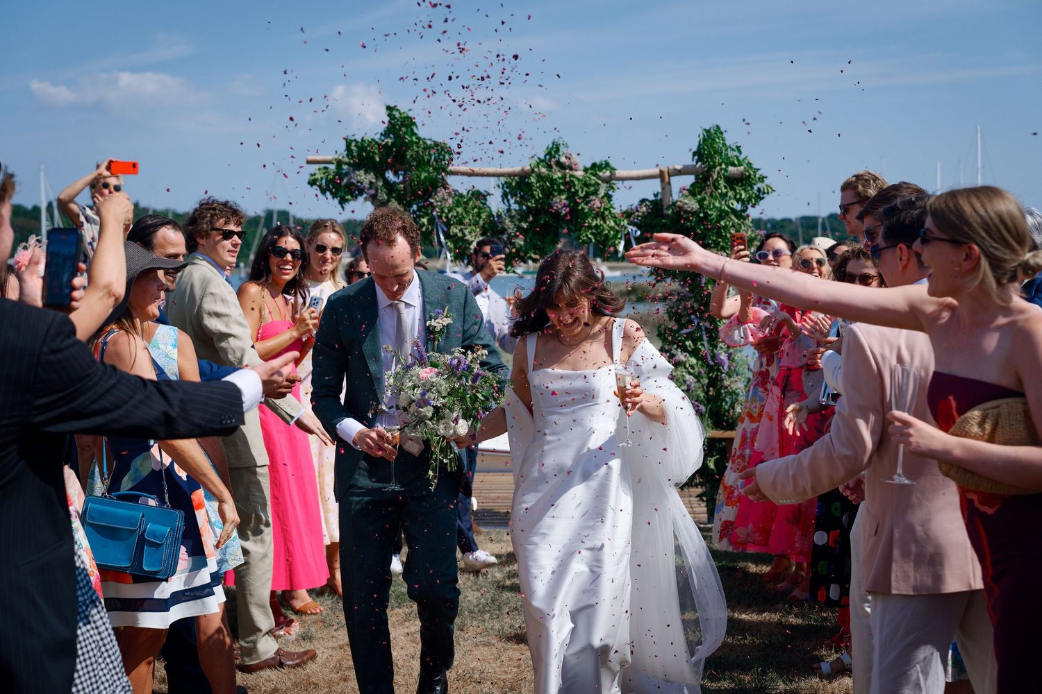 Bride and groom walk through confetti shower surrounded by cheering wedding guests outdoors on sunny day.