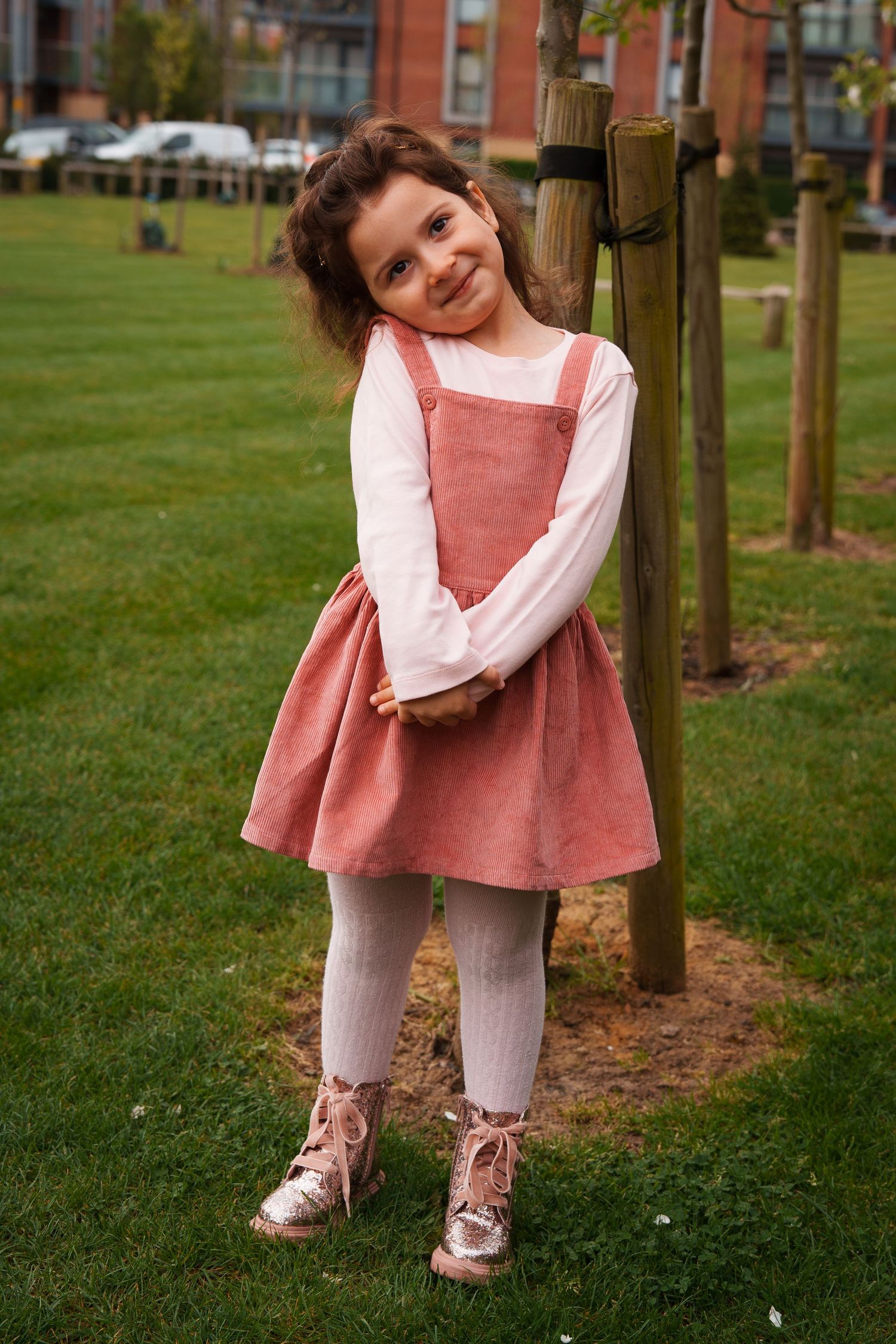 Young girl in pink dress and white long-sleeve shirt smiling while leaning against wooden post in grassy park area.